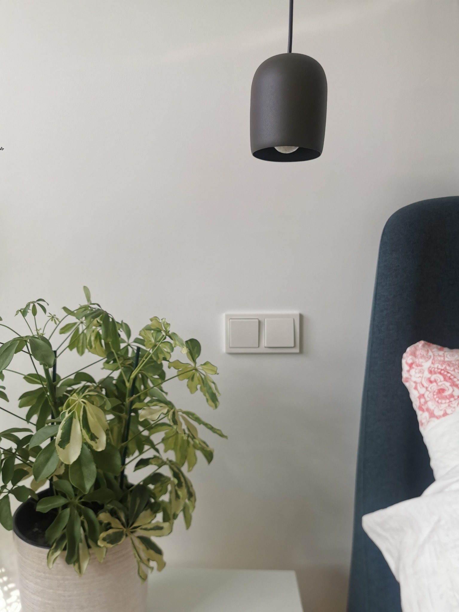 Minimalistyczne wnętrze sypialni: wisząca lampa w kształcie walca, podwójny włącznik światła, roślina doniczkowa z zielono-żółtymi liśćmi oraz zagłówek łóżka z poduszką w tle.
