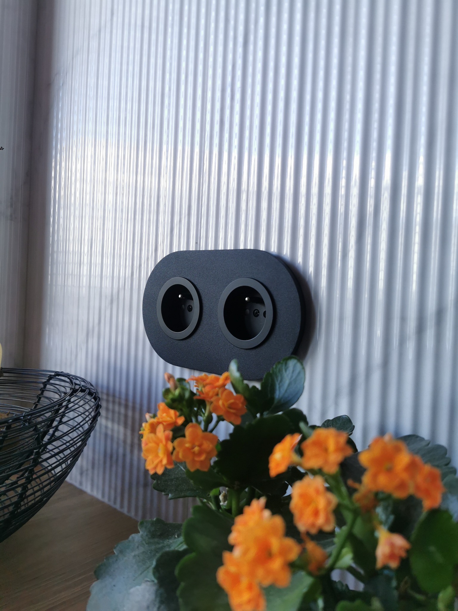 Czarny, podwójny kontakt elektryczny z dekoracyjną ramką na tle białej, żłobkowanej ściany, z pomarańczowymi kwiatami w doniczce w dolnej części kadru.