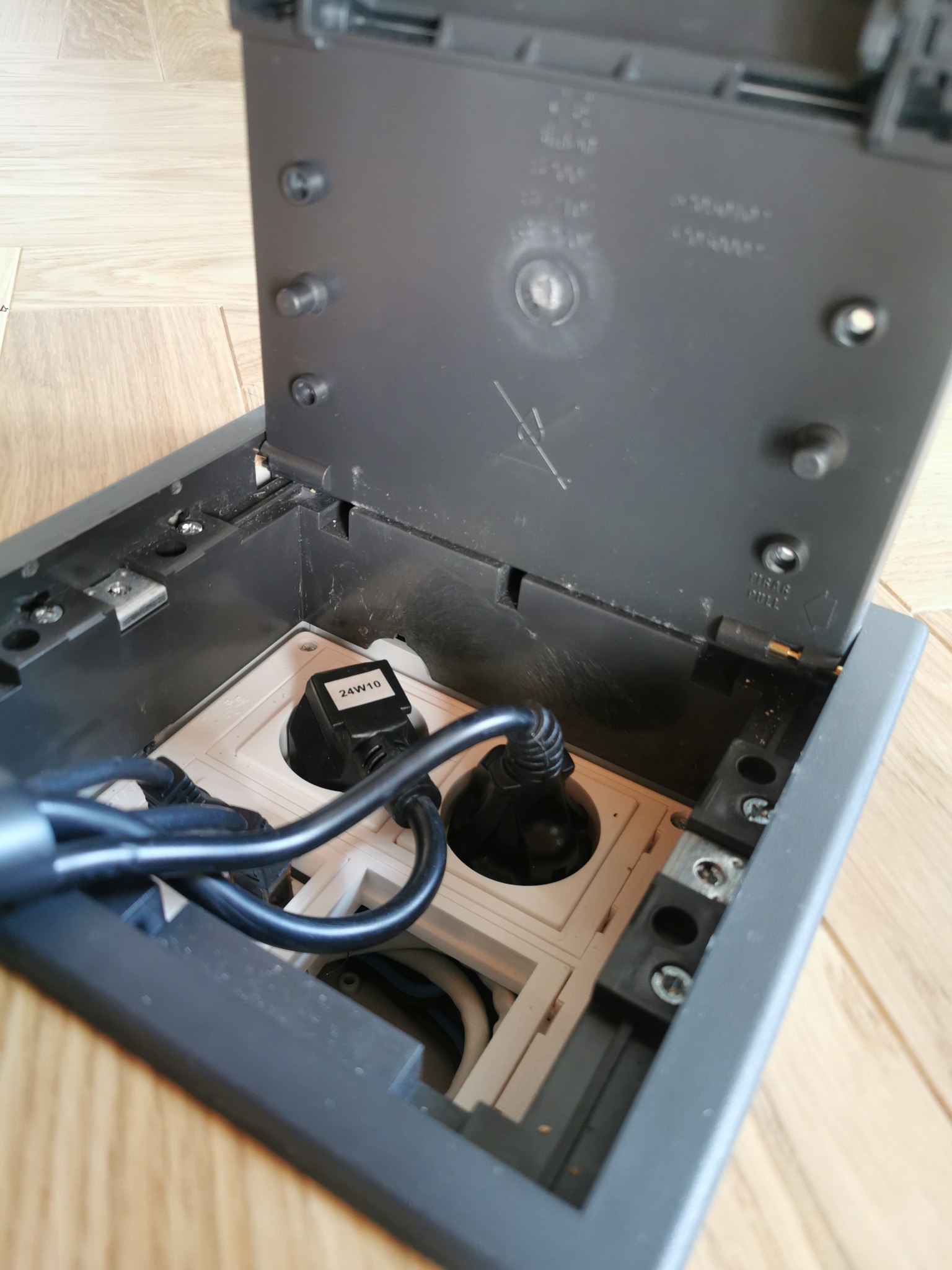 Otwarta podłogowa puszka elektryczna z dwoma gniazdami, wpięte kable zasilające, widoczne elementy montażowe i przewody wewnątrz.