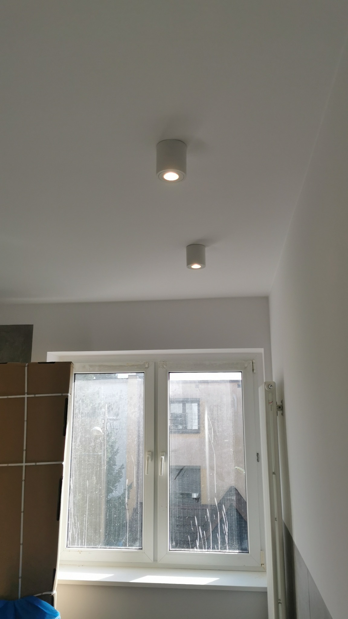 Dwa cylindryczne, białe reflektory sufitowe LED, świecące ciepłym światłem, zamontowane na białym suficie w nowym mieszkaniu z brudnymi oknami.