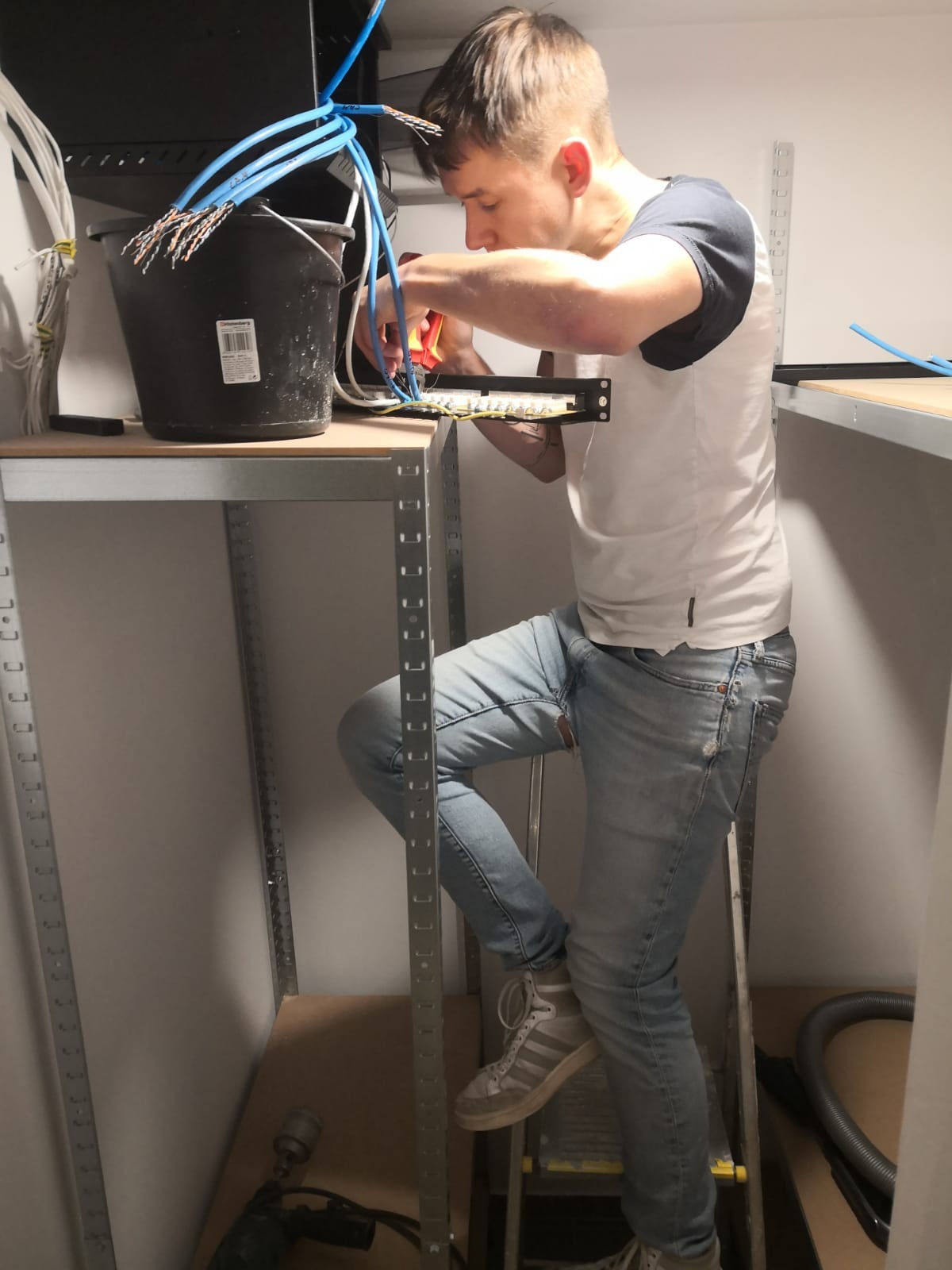 Młody mężczyzna na drabinie, podłącza kable telekomunikacyjne do panelu krosowniczego, używając zaciskarki. Wiązka kabli w czarnym wiadrze obok.
