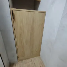 Zamknięta szafka z drewnianymi drzwiczkami, z czarnym uchwytem, wkomponowana w jasną, marmurkową ścianę i podłogę. Ujęcie z dołu.