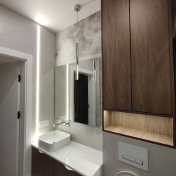 Paweł Kmieć Meble przy lesie - Nowoczesna łazienka z drewnianą szafką nad toaletą, umywalką na blacie i podświetlanym lustrem. Minimalistyczny design, ciepłe oświetlenie.