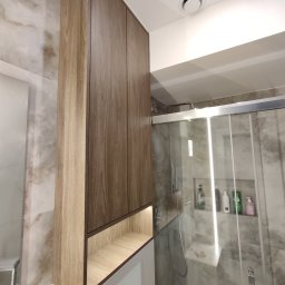 Paweł Kmieć Meble przy lesie - Wysoka, drewniana szafka łazienkowa z podświetlaną półką nad toaletą, w tle kabina prysznicowa z półkami na kosmetyki. Nowoczesny design, jasne płytki.