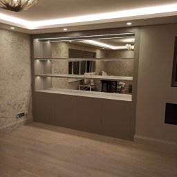 Paweł Kmieć Meble przy lesie - Szara zabudowa meblowa z lustrem i oświetleniem LED w salonie. Widoczne półki i szafki. W lustrze odbija się biurko i komputer. Parkiet na podłodze.