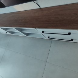 Nowoczesna szafka RTV z drewnianym blatem i białymi frontami szuflad z czarnymi uchwytami, ustawiona na jasnych płytkach podłogowych. Widok z góry.
