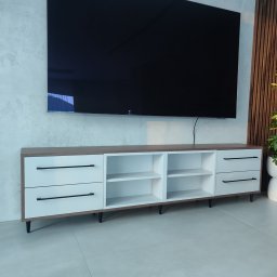 Nowoczesna szafka RTV z białymi frontami i czarnymi uchwytami, umieszczona pod telewizorem na tle ściany z dekoracyjnymi panelami drewnianymi.