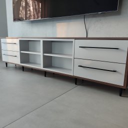 Nowoczesna szafka RTV z białymi frontami i drewnianym korpusem, czarne uchwyty, ustawiona na tle szarej ściany z telewizorem. Minimalistyczny design.