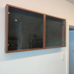 Dwie przeszklone szafki wiszące z brązowymi ramami na białej ścianie, jedna otwarta, ukazująca zawartość. Widoczny włącznik światła i fragment otwartego przejścia.