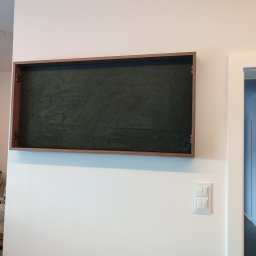 Paweł Kmieć Meble przy lesie - Otwarta szafka wnękowa z ciemnozielonym, welurowym wnętrzem i drewnianą ramą, zamontowana na białej ścianie obok włącznika światła i drzwi.