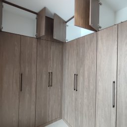 Paweł Kmieć Meble przy lesie - Jasna, narożna szafa wnękowa z drewnianymi frontami i czarnymi uchwytami. Część szafek jest otwarta, ukazując białe wnętrze. Widok z dołu, oświetlenie górne.