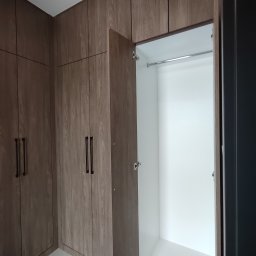 Paweł Kmieć Meble przy lesie - Wnętrze szafy wnękowej z otwartymi drzwiami, widoczny drążek na ubrania. Fronty szafy w odcieniach brązu z wyraźnym rysunkiem drewna, ciemne, podłużne uchwyty.