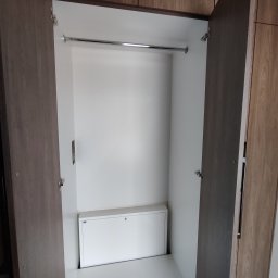 Paweł Kmieć Meble przy lesie - Otwarta szafa wnękowa z drążkiem i białą szafką w dolnej części. Widoczne zawiasy i faktura drewna na drzwiach. Minimalistyczny design.