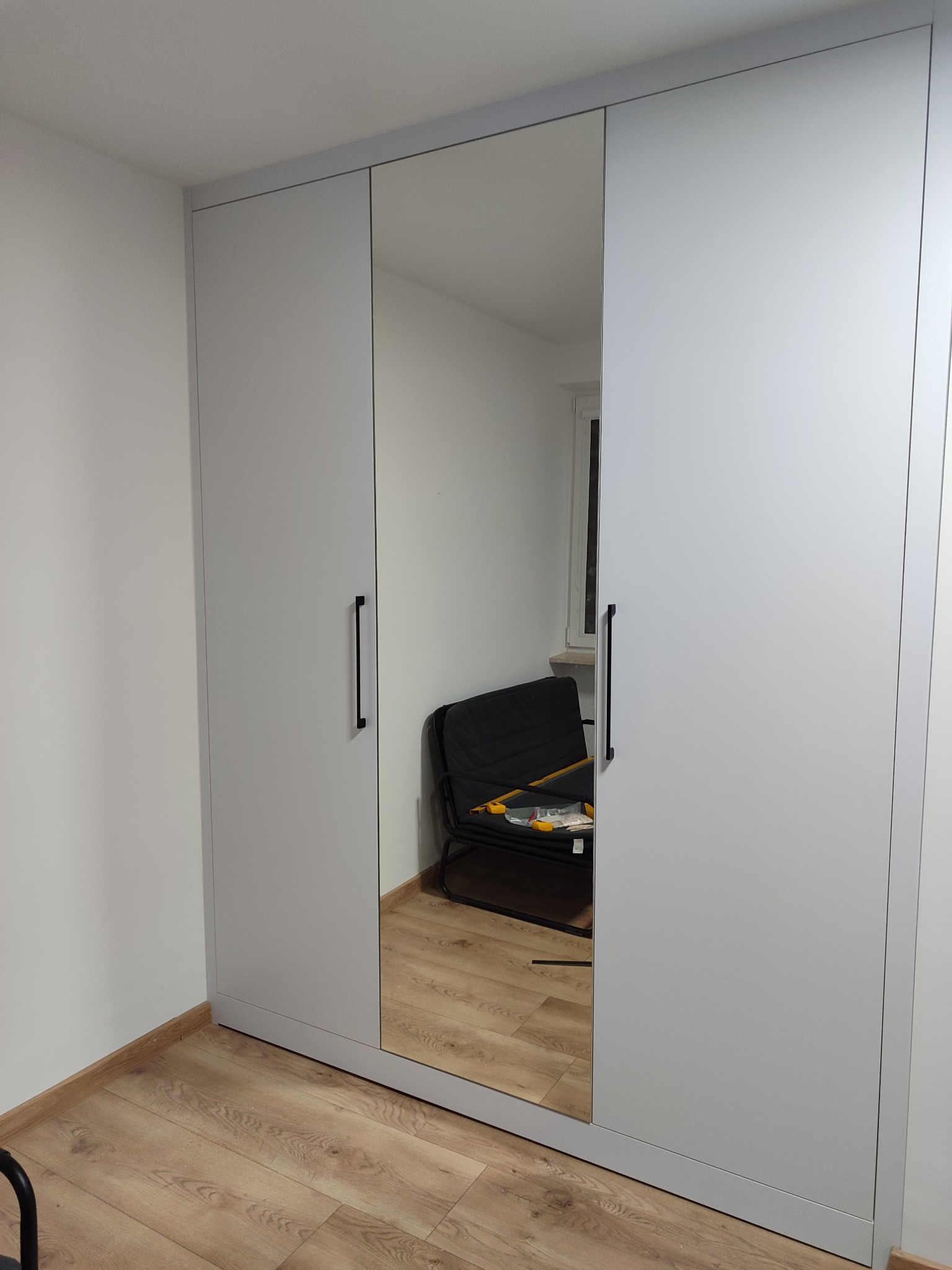 Jasnoszara szafa trzydrzwiowa z lustrem pośrodku, z długimi, pionowymi uchwytami, na tle parkietu i białych ścian. W lustrze odbija się krzesło.