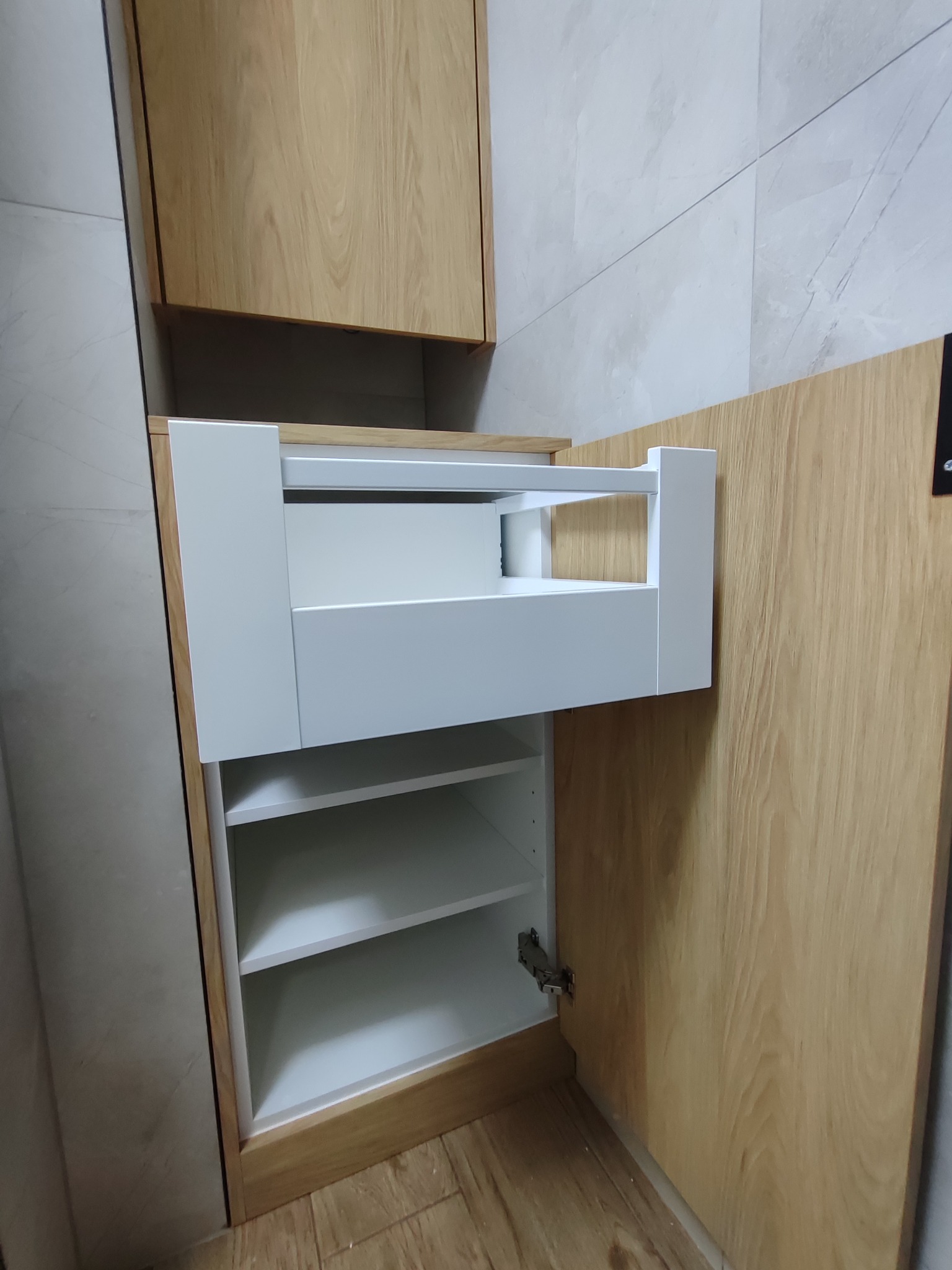 Szafka łazienkowa z jasnego drewna z otwartą białą szufladą i półkami wewnątrz. Widoczne zawiasy i marmurowe płytki na ścianie w tle.