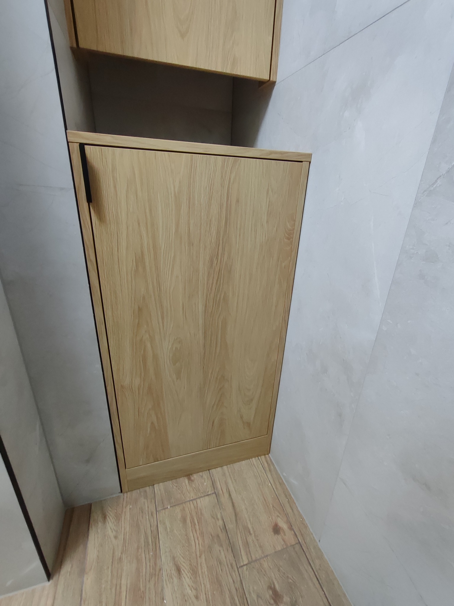 Zamknięta szafka z drewnianymi drzwiczkami, z czarnym uchwytem, wkomponowana w jasną, marmurkową ścianę i podłogę. Ujęcie z dołu.