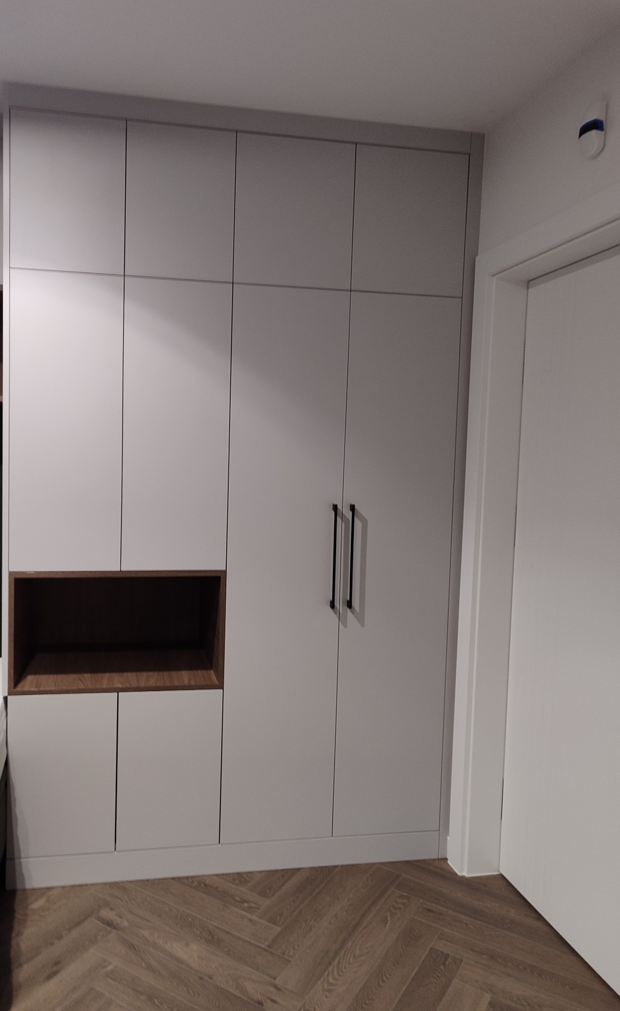 Biała szafa wnękowa z ciemnym, drewnianym regałem, minimalistyczne uchwyty, parkiet w jodełkę, widok na fragment drzwi. Nowoczesny design, stonowana kolorystyka.