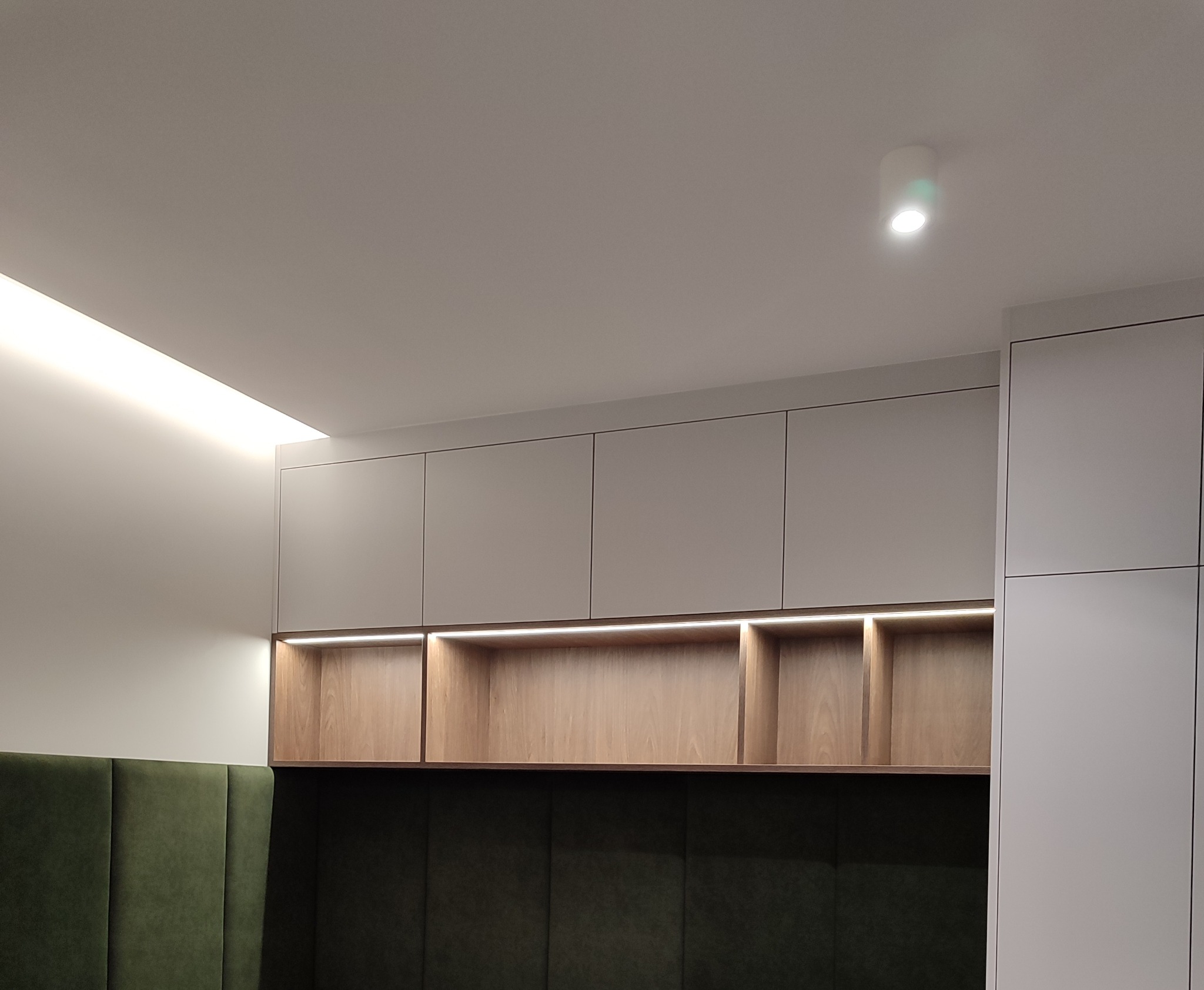 Nowoczesna zabudowa meblowa z podświetlanymi półkami i tapicerowanym panelem ściennym w odcieniach szarości, drewna i zieleni. Minimalistyczny design, oświetlenie LED.