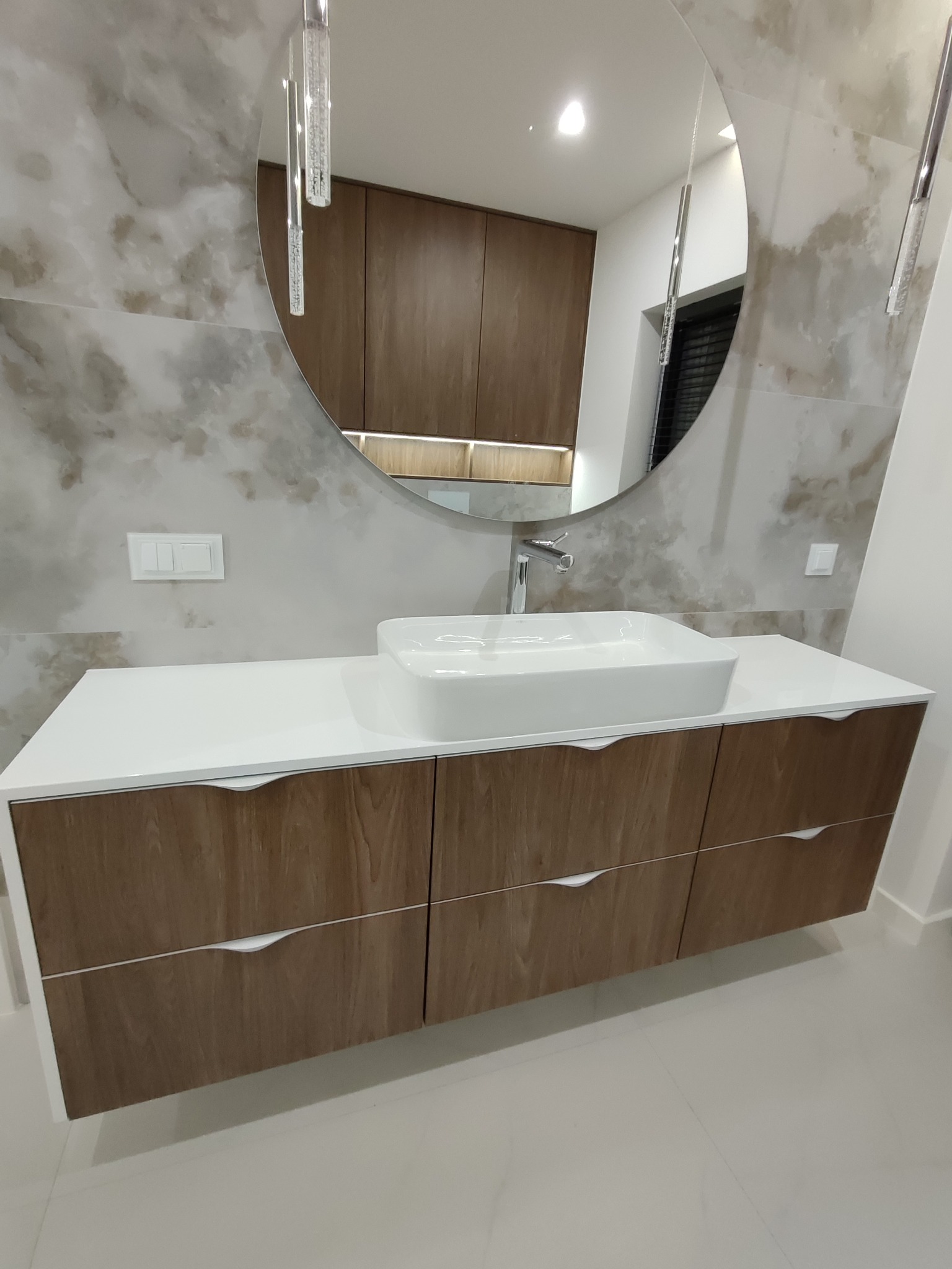 Nowoczesna szafka łazienkowa z umywalką nablatową i dużym okrągłym lustrem. Fronty szafek w odcieniach drewna, biały blat i marmurowe wykończenie ściany.