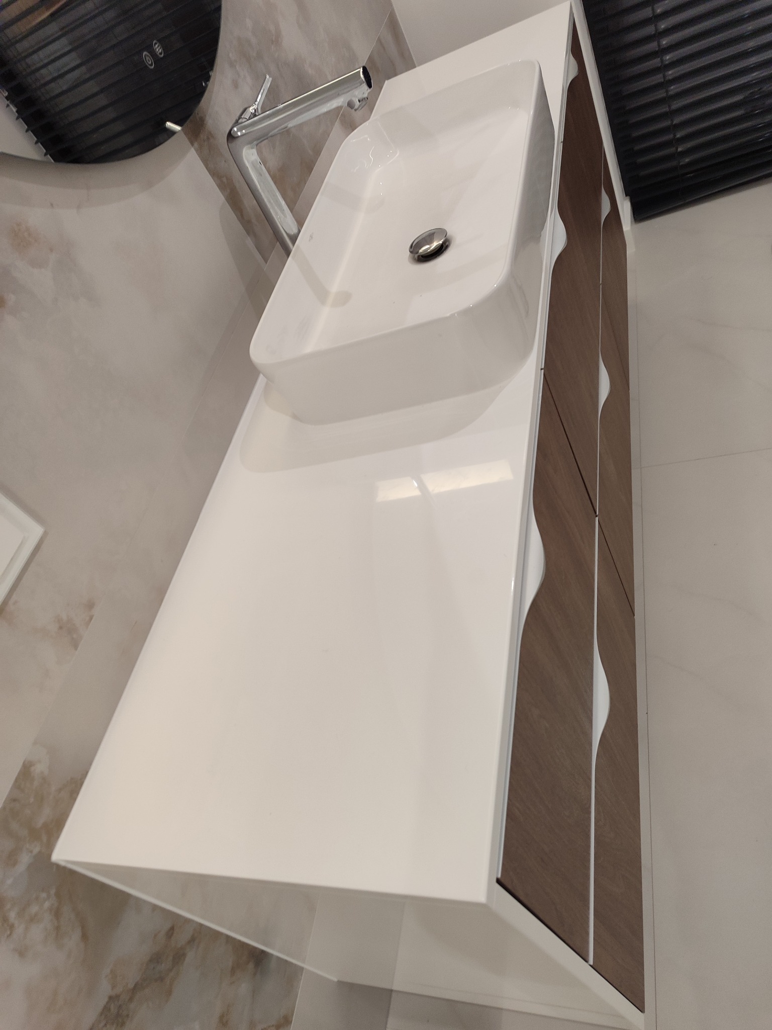 Nowoczesna szafka łazienkowa z umywalką nablatową i chromowaną baterią. Fronty szafki w kolorze drewna z białymi, falistymi wstawkami. Minimalistyczny design.