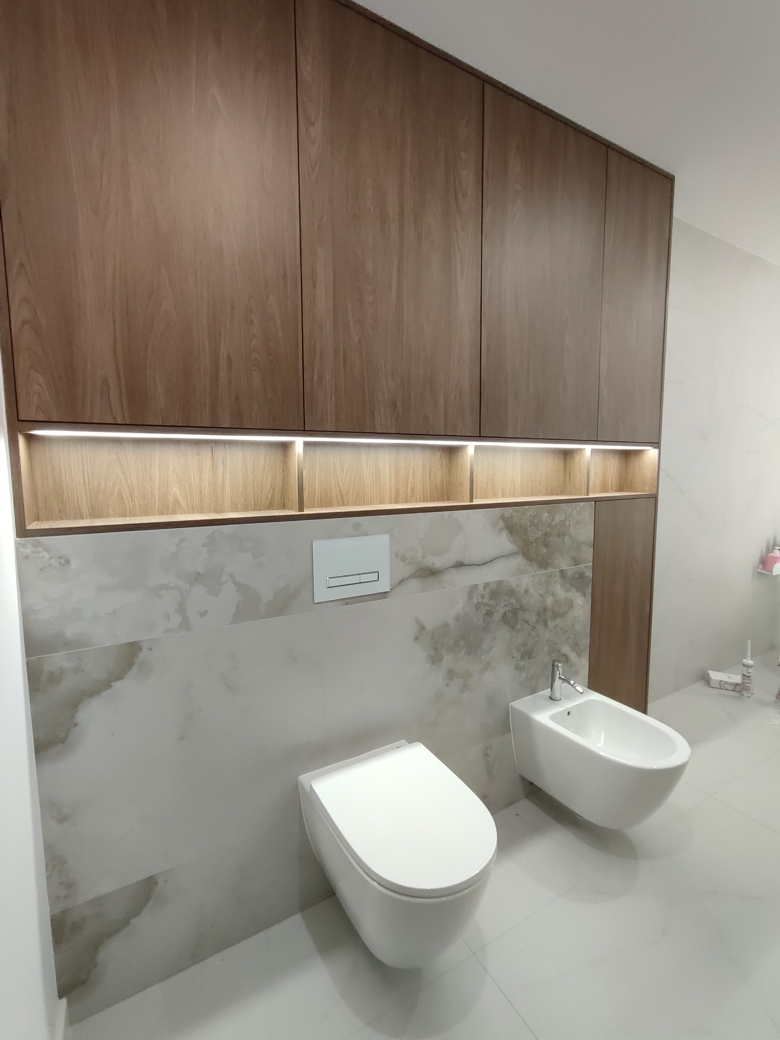 Nowoczesna łazienka z zabudową meblową z drewna, podświetlanymi półkami i marmurowymi płytkami. Toaleta i bidet w minimalistycznym stylu.