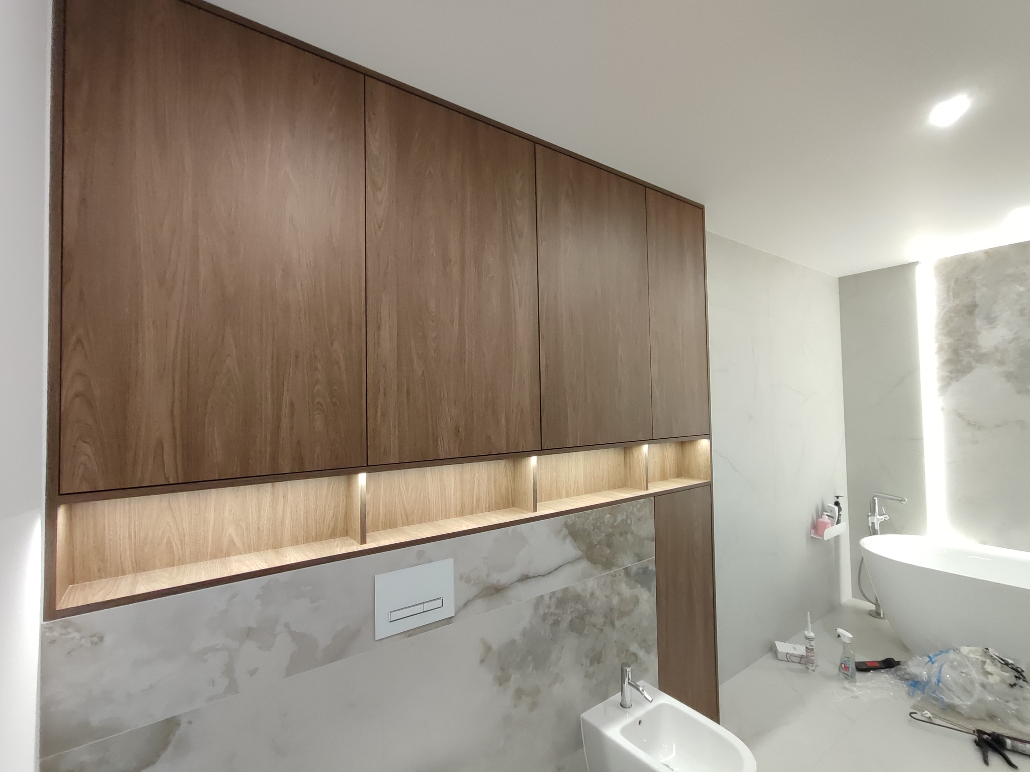 Drewniane szafki łazienkowe z podświetlanymi półkami, umywalka i bidet na tle marmurowych płytek. Widoczne akcesoria łazienkowe i narzędzia montażowe.