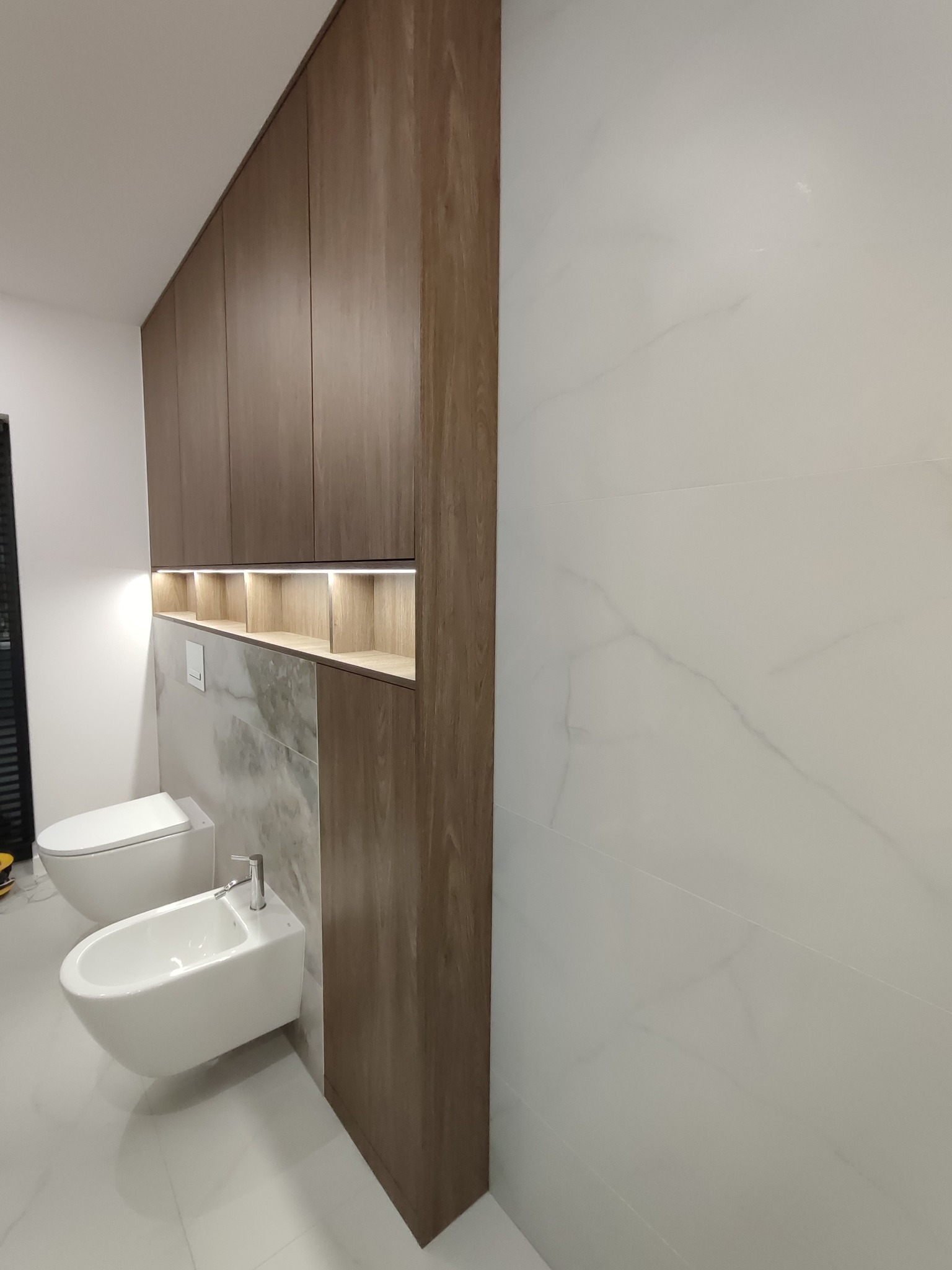 Nowoczesna łazienka z zabudową meblową z drewna, podświetlane półki nad toaletą i bidetem, białe marmurowe płytki na ścianach i podłodze. Minimalistyczny design.