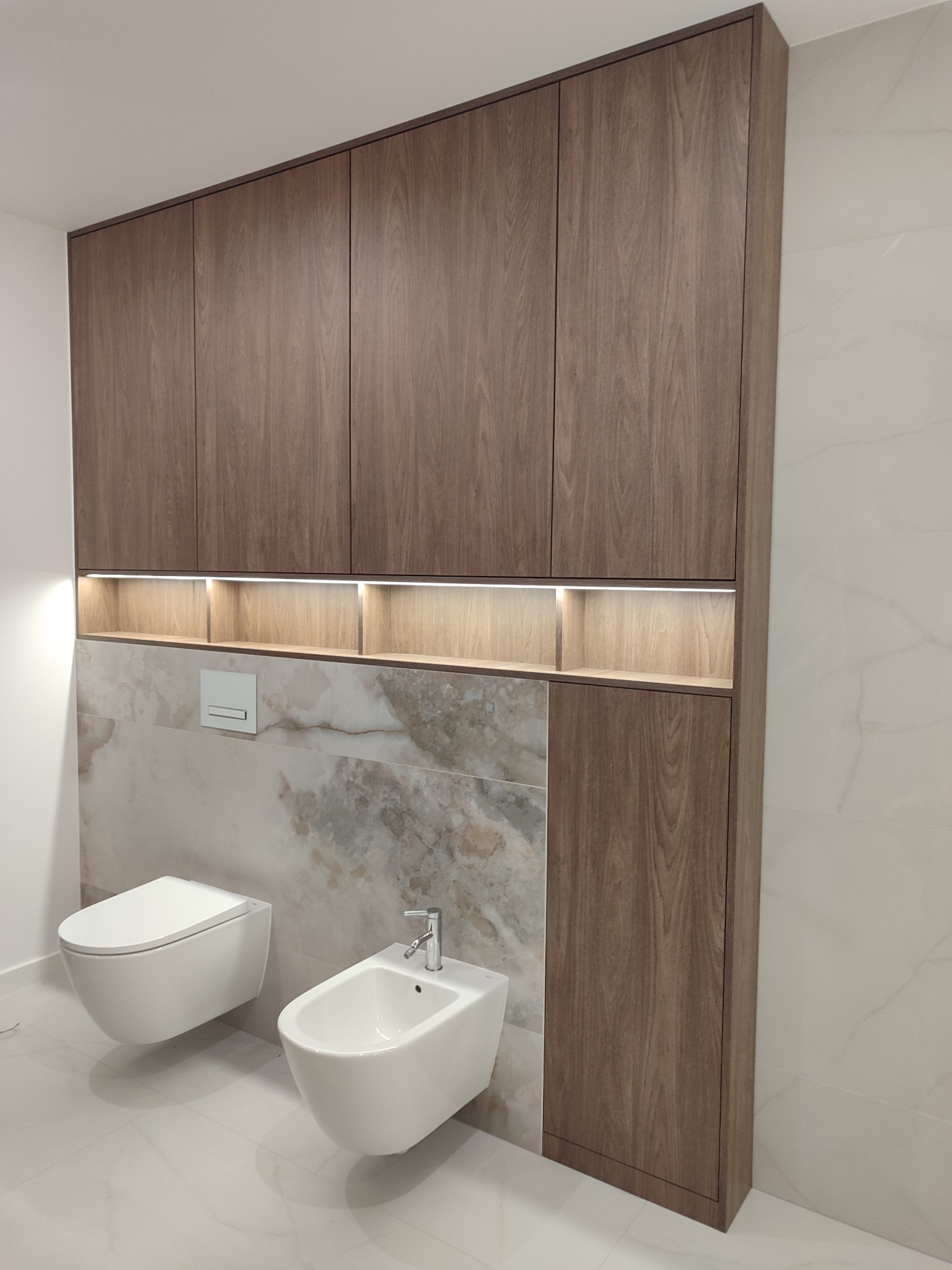 Nowoczesna zabudowa łazienkowa z szafkami z drewna orzechowego, podświetlanymi półkami i marmurowym panelem nad toaletą i bidetem. Minimalistyczny design.
