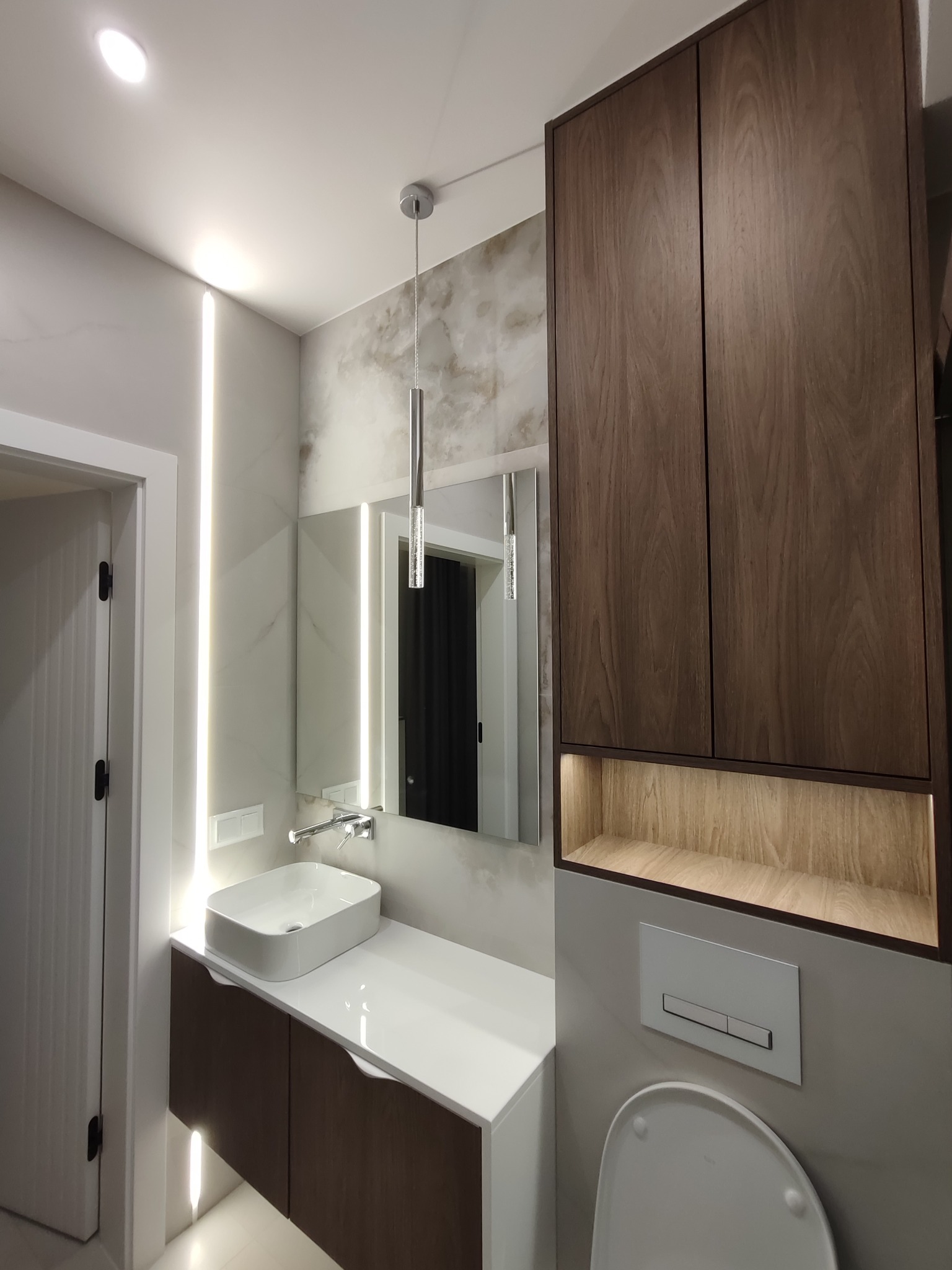 Nowoczesna łazienka z drewnianą szafką nad toaletą, umywalką na blacie i podświetlanym lustrem. Minimalistyczny design, ciepłe oświetlenie.