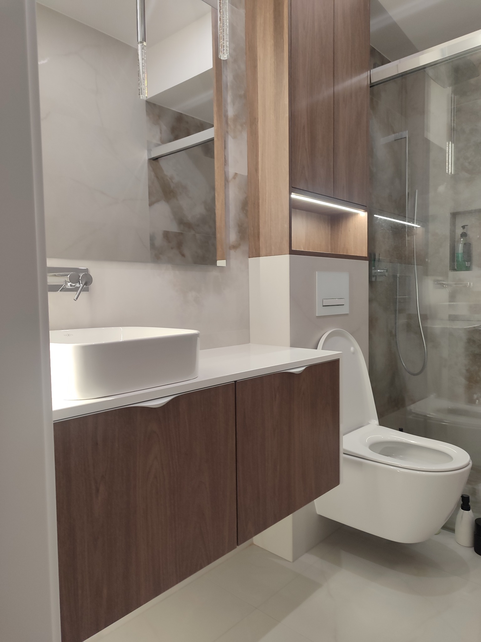 Nowoczesna łazienka z drewnianą szafką pod umywalką, wiszącą toaletą i kabiną prysznicową. Minimalistyczny design, ciepłe oświetlenie i marmurowe wykończenie ścian.