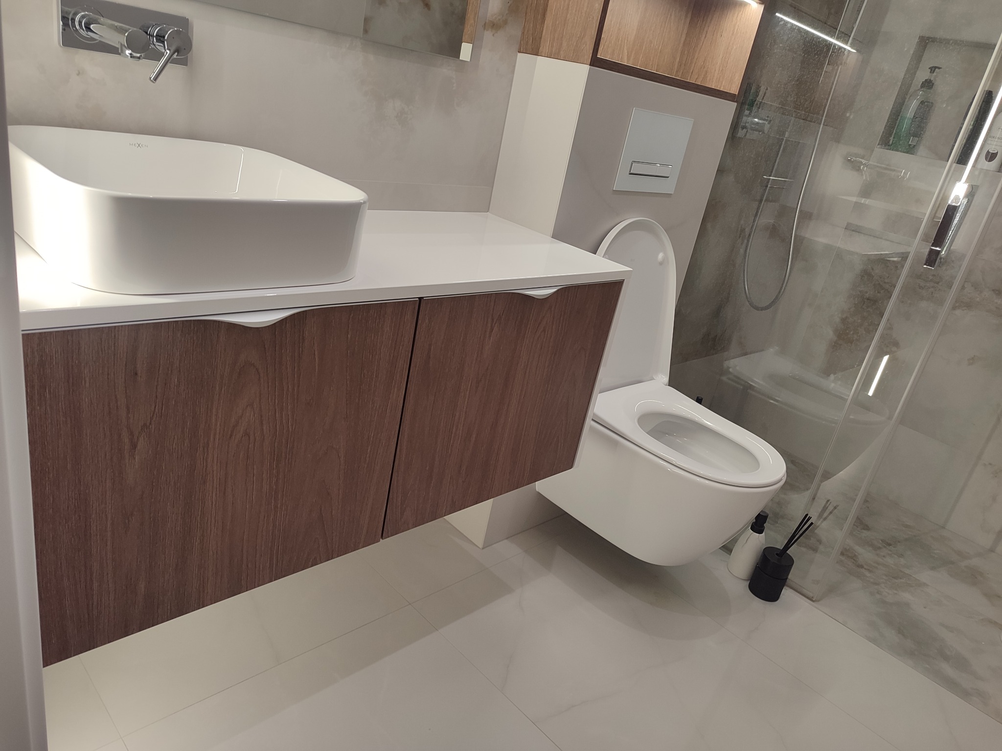 Nowoczesna łazienka z drewnianą szafką pod umywalką, białą ceramiką i kabiną prysznicową. Minimalistyczny design, jasne płytki podłogowe, eleganckie wykończenie.