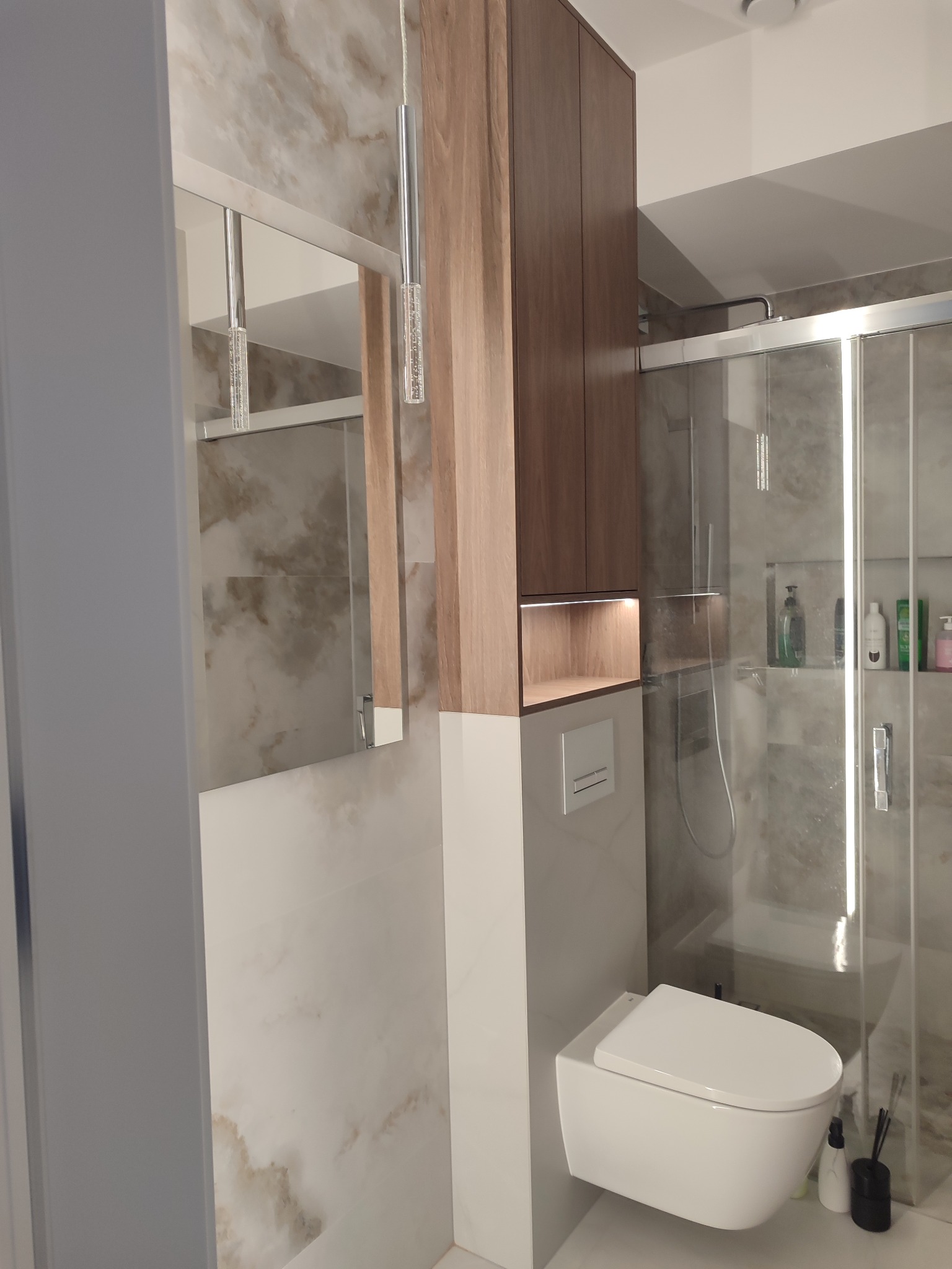 Nowoczesna łazienka z podwieszaną toaletą, szafką z oświetleniem LED i lustrem. Marmurowe płytki na ścianach i szklana kabina prysznicowa.