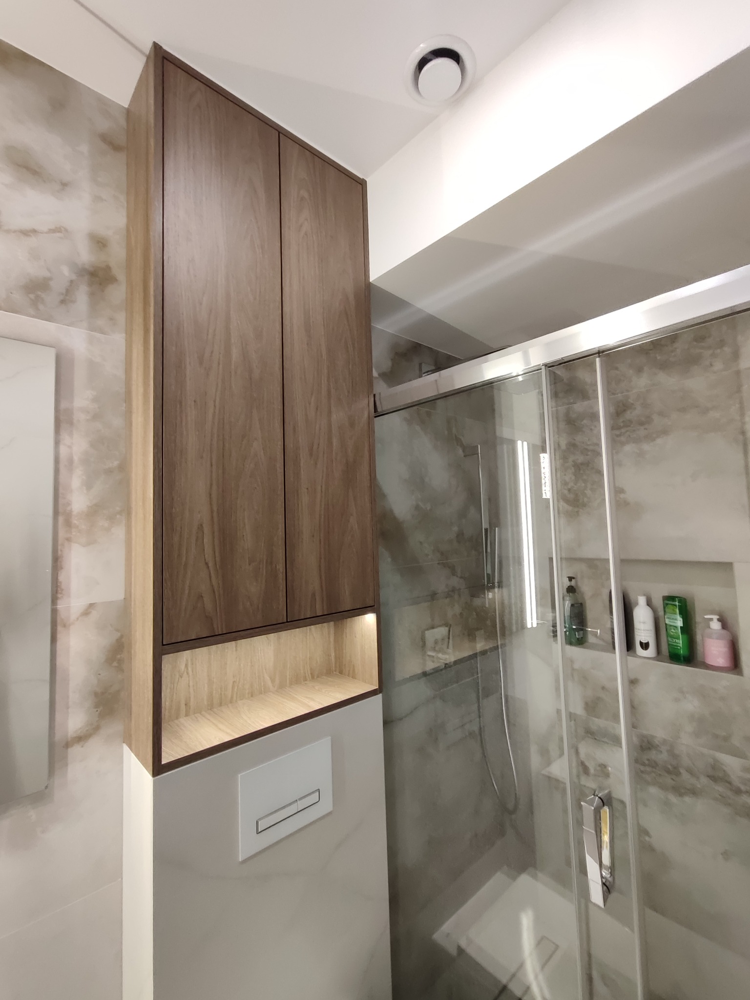 Wysoka, drewniana szafka łazienkowa z podświetlaną półką nad toaletą, obok kabiny prysznicowej z marmurowymi płytkami. Nowoczesny design, oświetlenie LED.