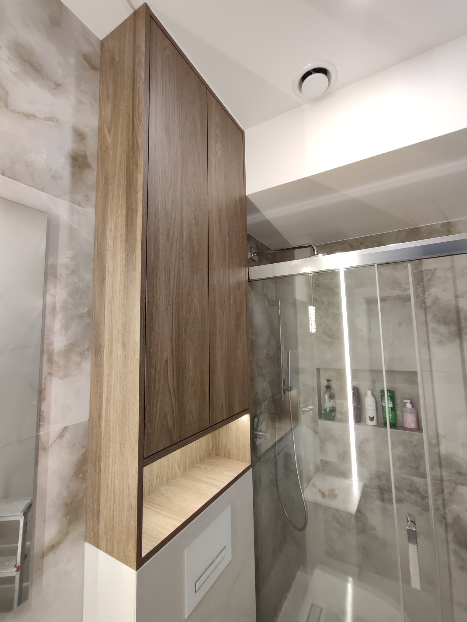 Wysoka, drewniana szafka łazienkowa z podświetlaną półką nad toaletą, w tle kabina prysznicowa z półkami na kosmetyki. Nowoczesny design, jasne płytki.