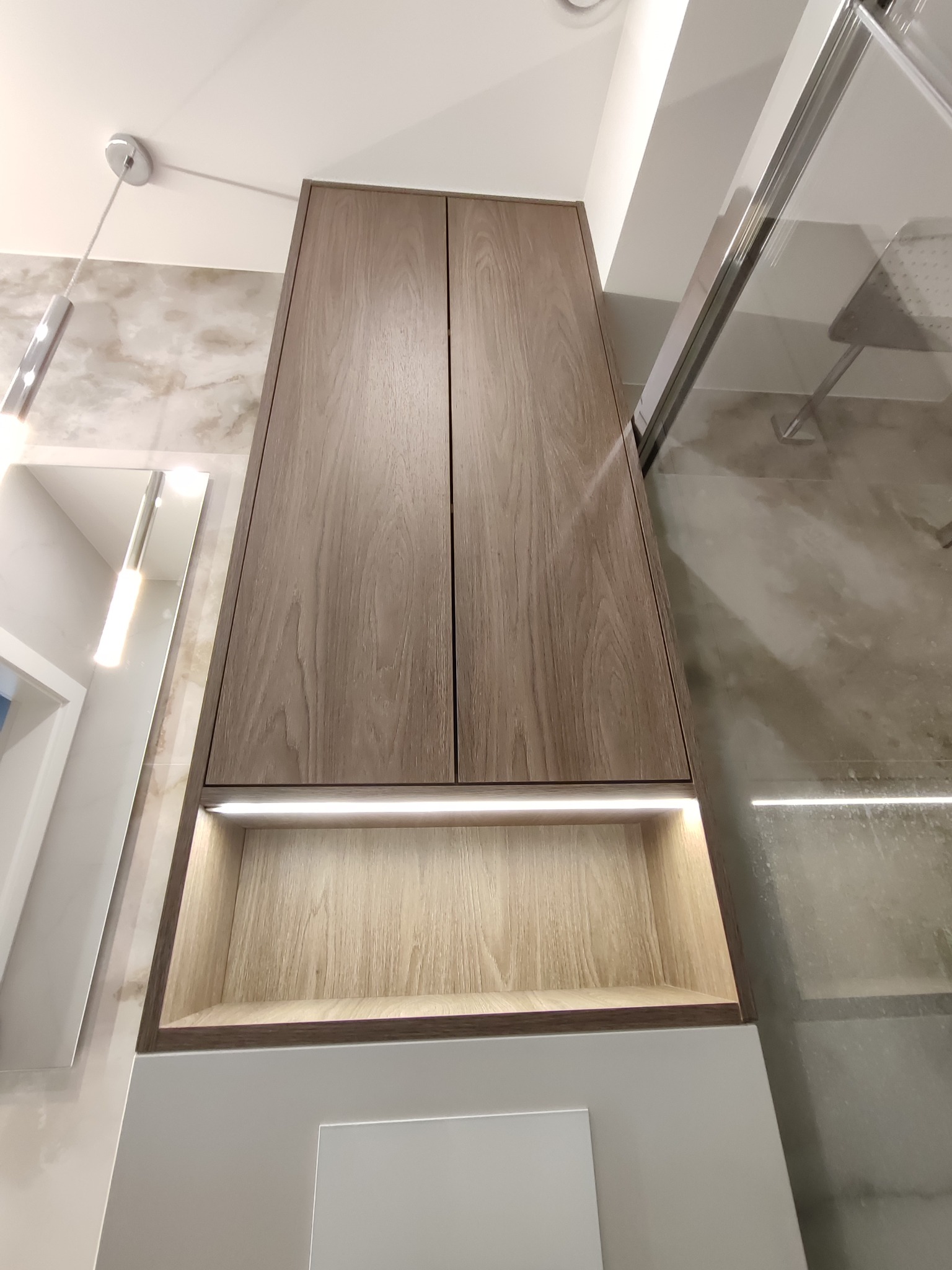 Drewniana szafka łazienkowa z oświetleniem LED, widok z dołu, obok kabina prysznicowa i lustro. Nowoczesny design, jasne wnętrze, minimalistyczny styl.