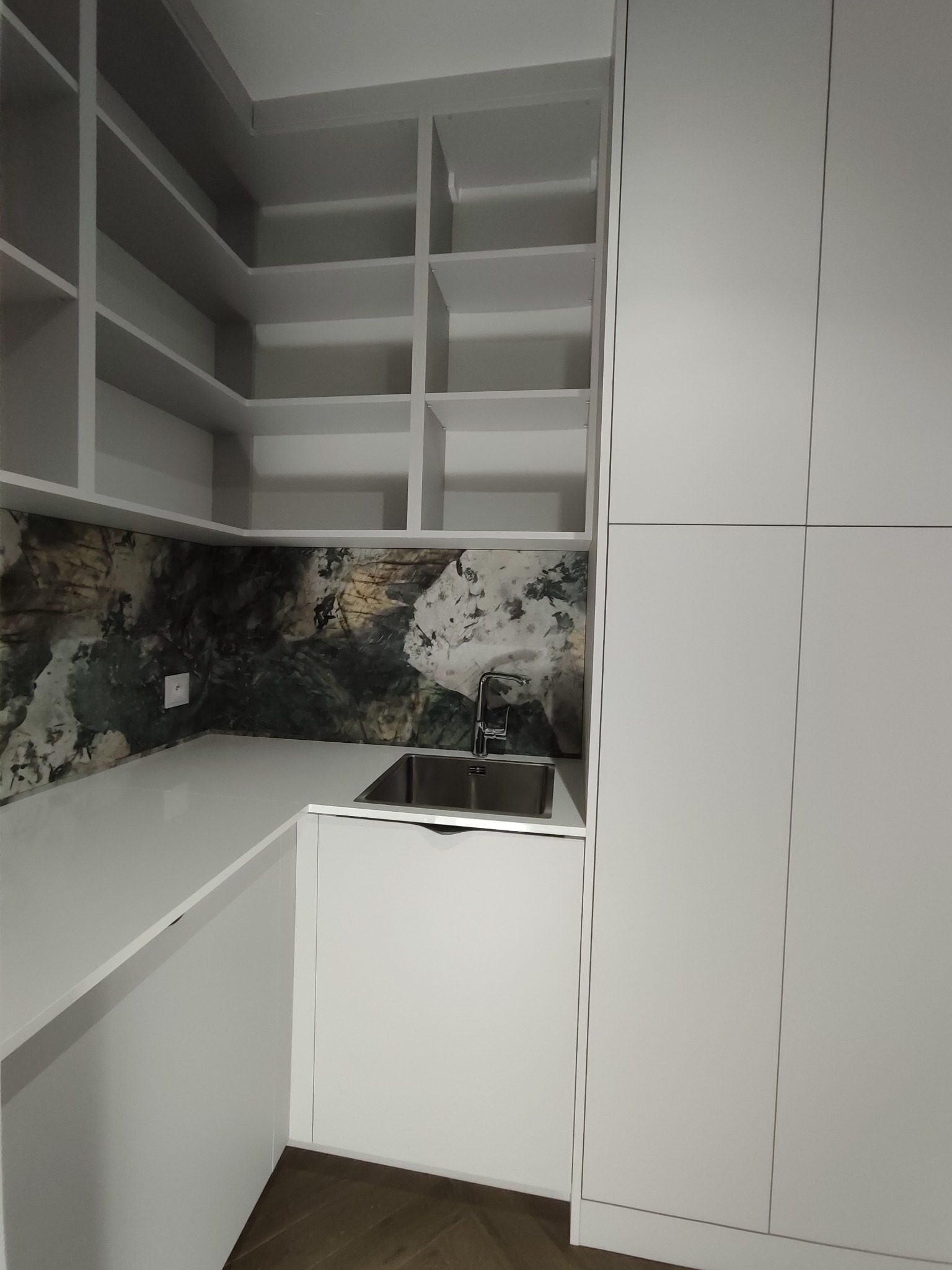 Biała zabudowa kuchenna z szafkami, półkami i blatem z umywalką. Na ścianie dekoracyjny panel imitujący marmur. Minimalistyczny design, nowoczesne wykończenie.