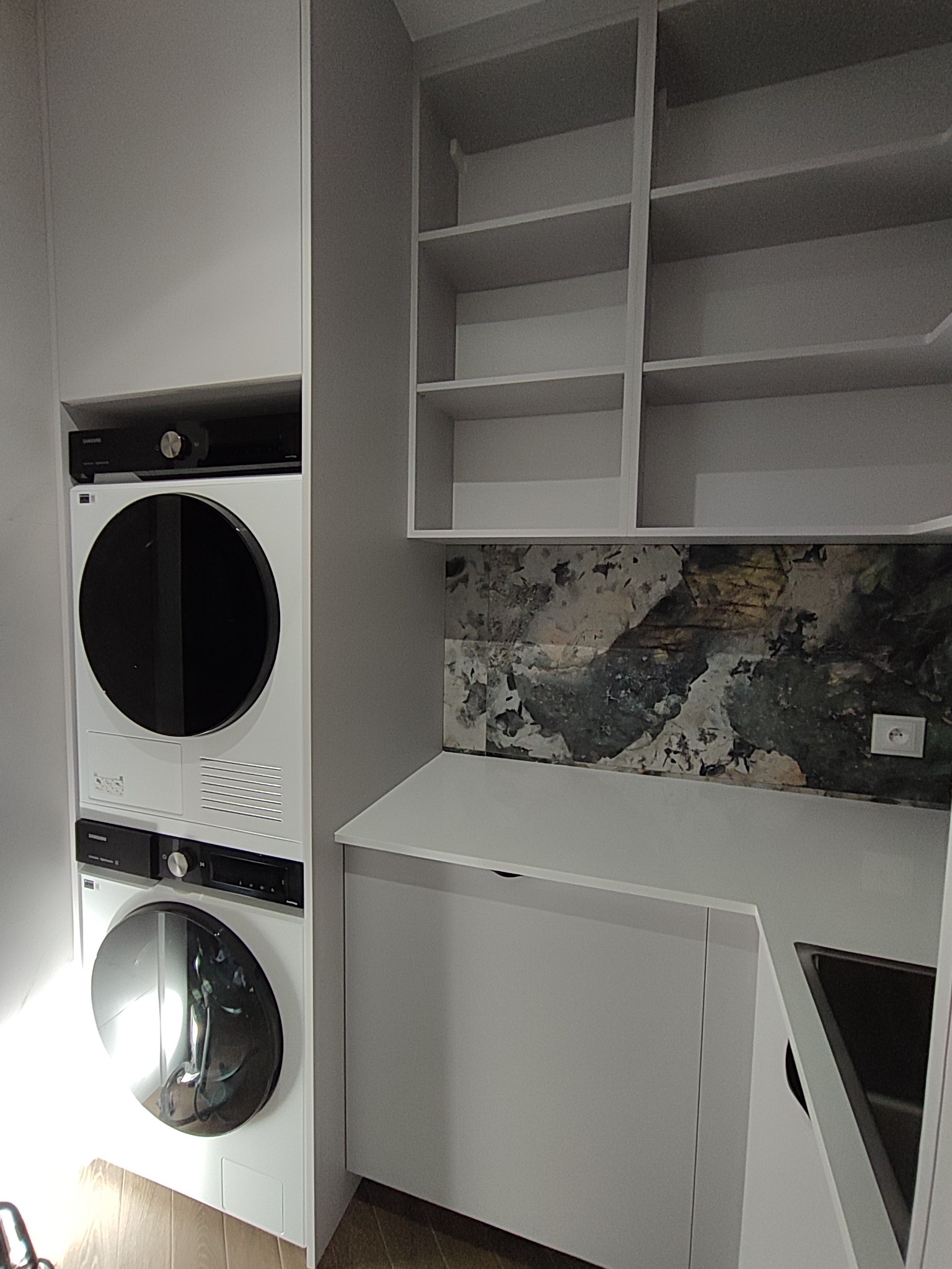 Biała zabudowa kuchenna z pralką i suszarką w kolumnie, otwarte półki i dekoracyjny panel nad blatem. Minimalistyczny design, nowoczesne wykończenie.