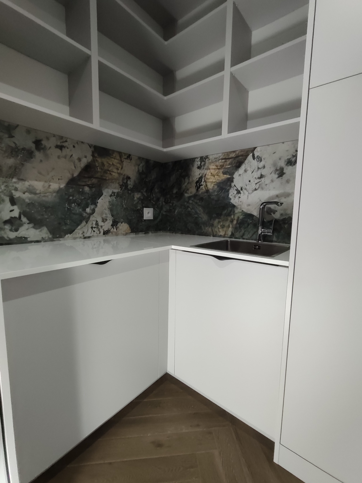 Nowoczesna, biała kuchnia na wymiar z marmurowym blatem i oryginalnym panelem ściennym. Widoczny zlew z baterią i otwarte półki.