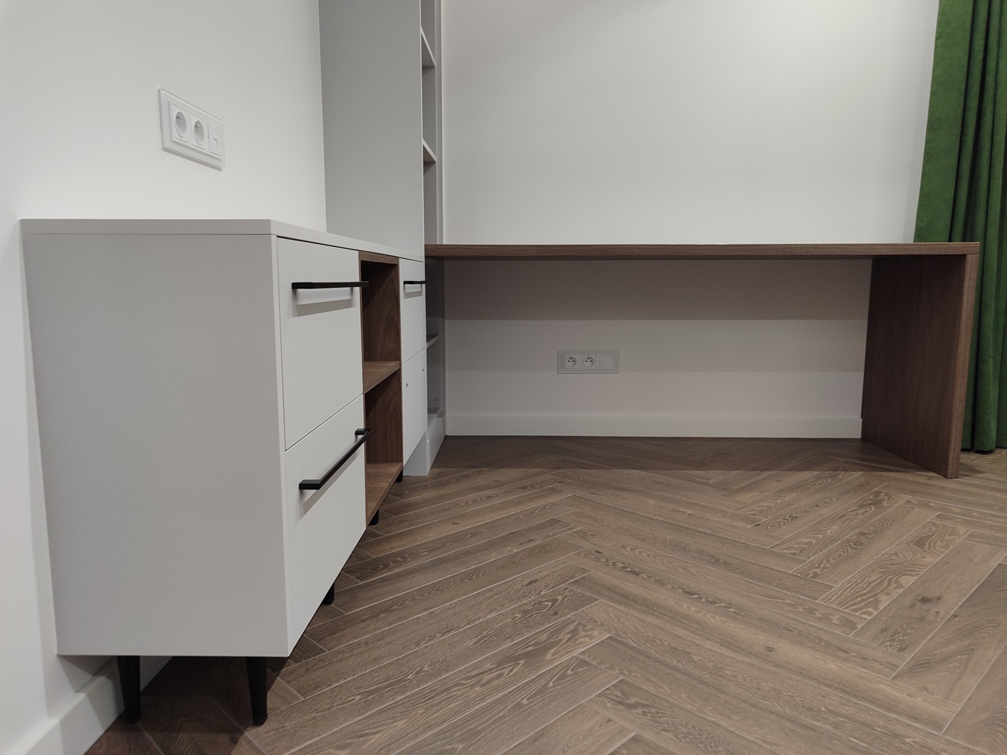 Nowoczesny zestaw mebli: biała komoda z czarnymi uchwytami, regał i biurko z drewnianym blatem, na parkiecie ułożonym w jodełkę. Minimalistyczny design, gniazdka w ścianie.