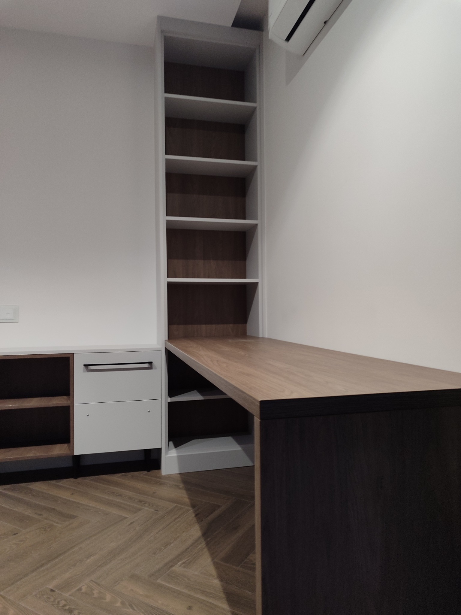 Nowoczesne biurko zintegrowane z regałem i szafkami w odcieniach bieli i drewna, na tle jasnej ściany i parkietu w jodełkę. Minimalistyczny design, oświetlenie górne.