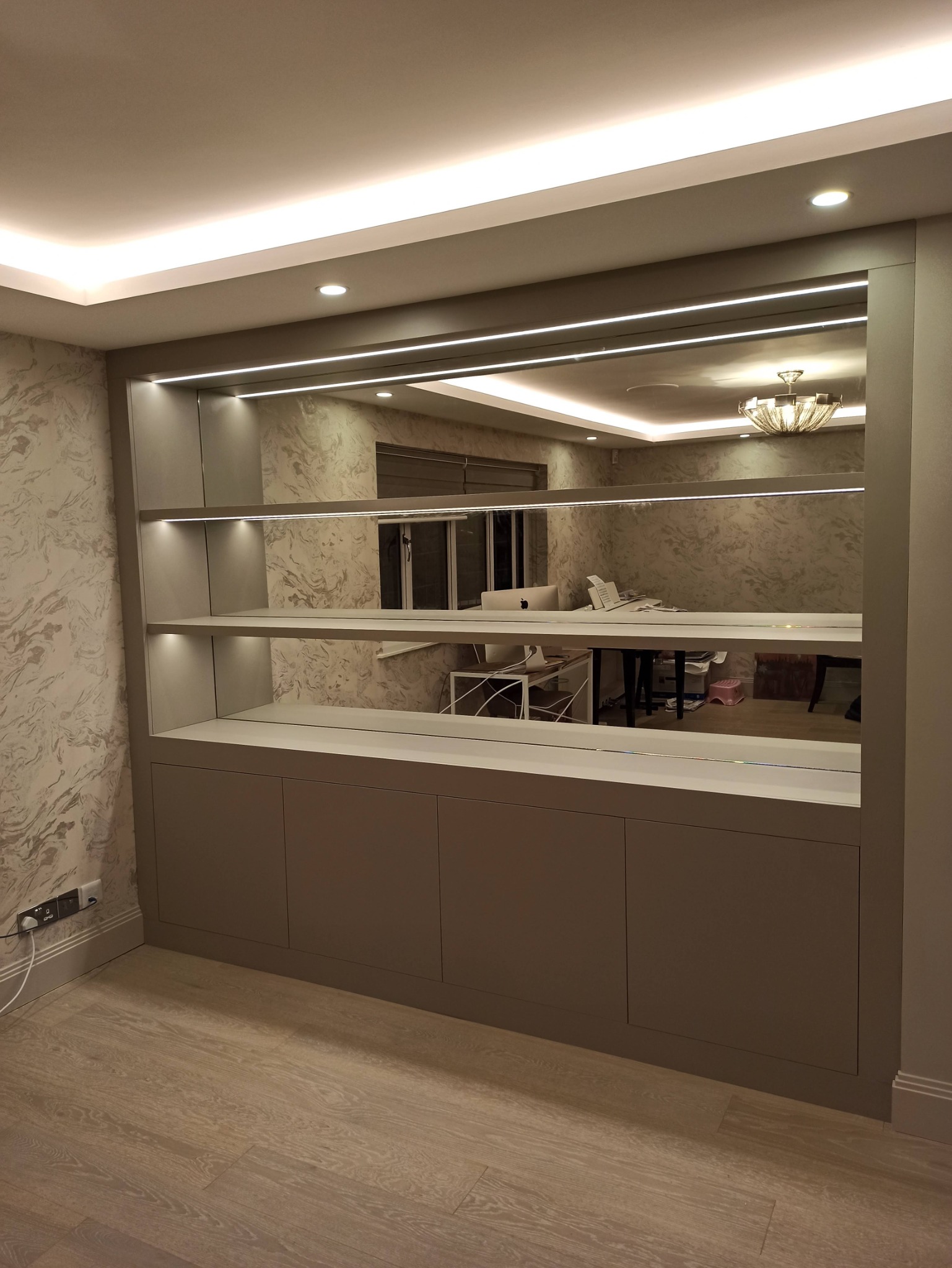 Szara zabudowa meblowa z szafkami i półkami, oświetlona listwami LED, z lustrzanymi plecami, odbijająca wnętrze pokoju z biurkiem i lampą.