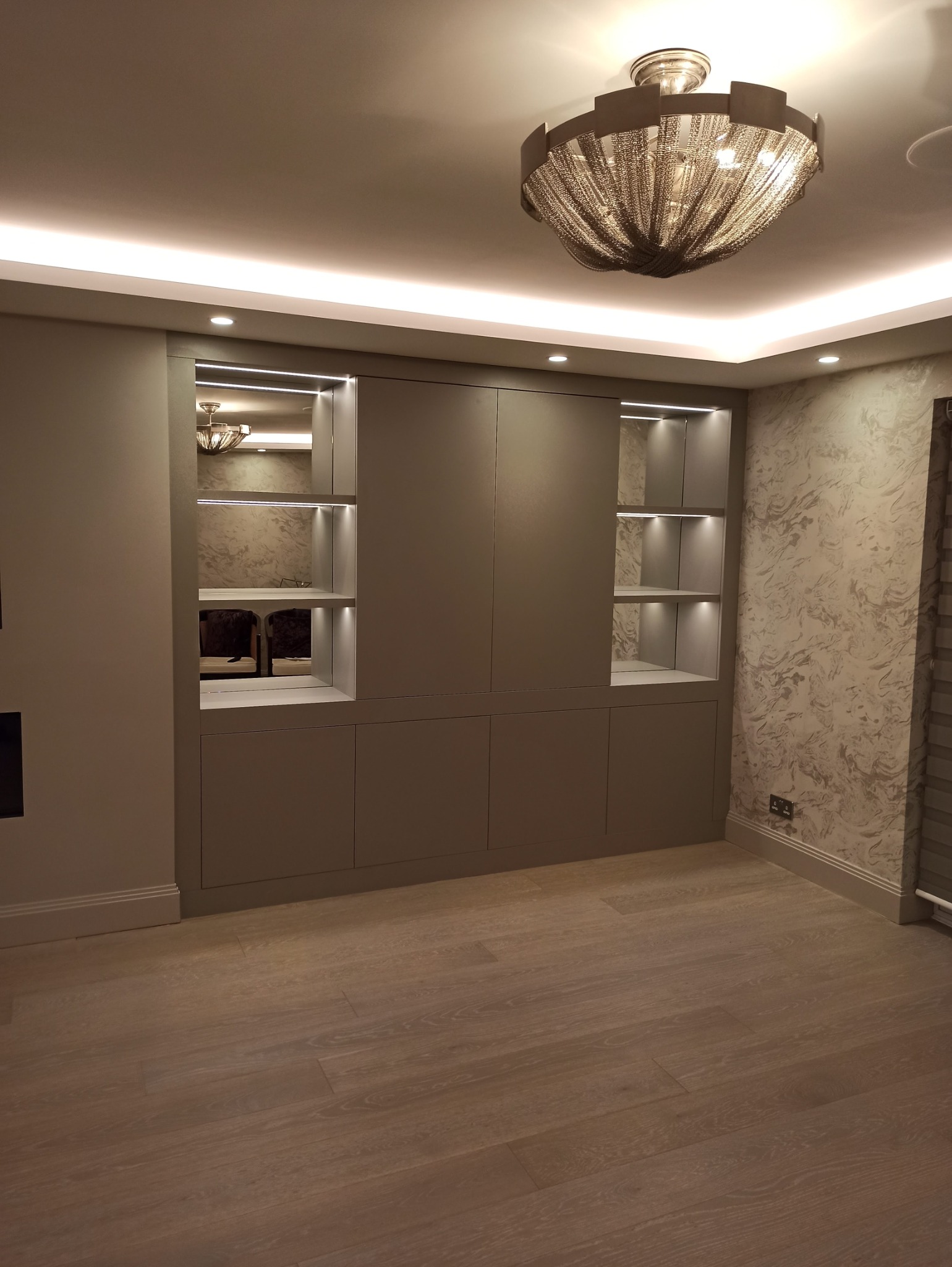 Elegancka, szara szafa wnękowa z podświetlanymi półkami, wkomponowana w ścianę. Widoczne oświetlenie sufitowe i parkiet.