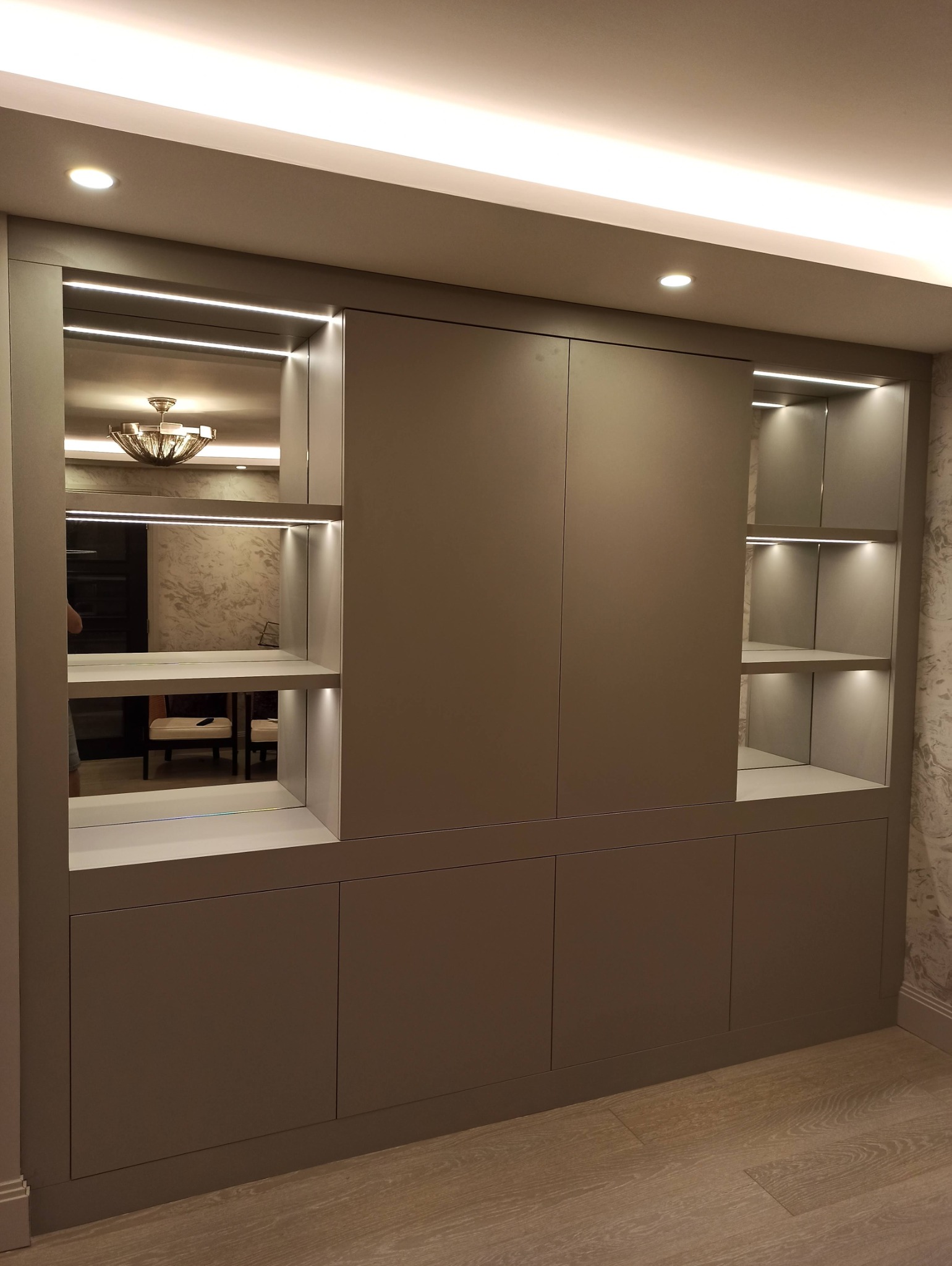 Szara zabudowa meblowa z oświetleniem LED, półkami i szafkami. W lustrzanych wnękach odbija się żyrandol, tworząc iluzję głębi. Minimalistyczny design.