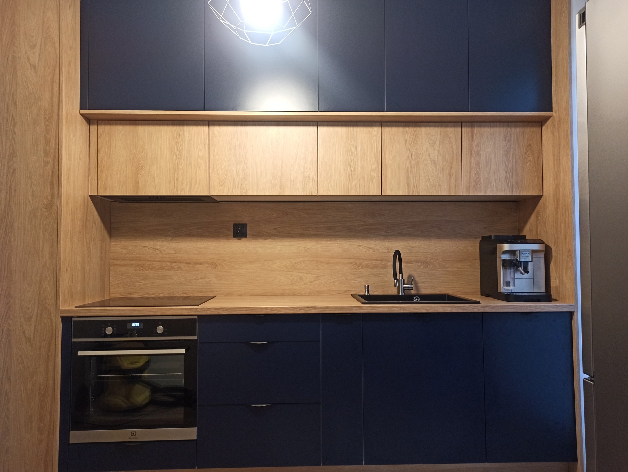 Nowoczesna kuchnia z ciemnoniebieskimi szafkami dolnymi i drewnianymi górnymi. Na blacie czarny zlew, ekspres do kawy i płyta indukcyjna. Nad blatem geometryczna lampa.