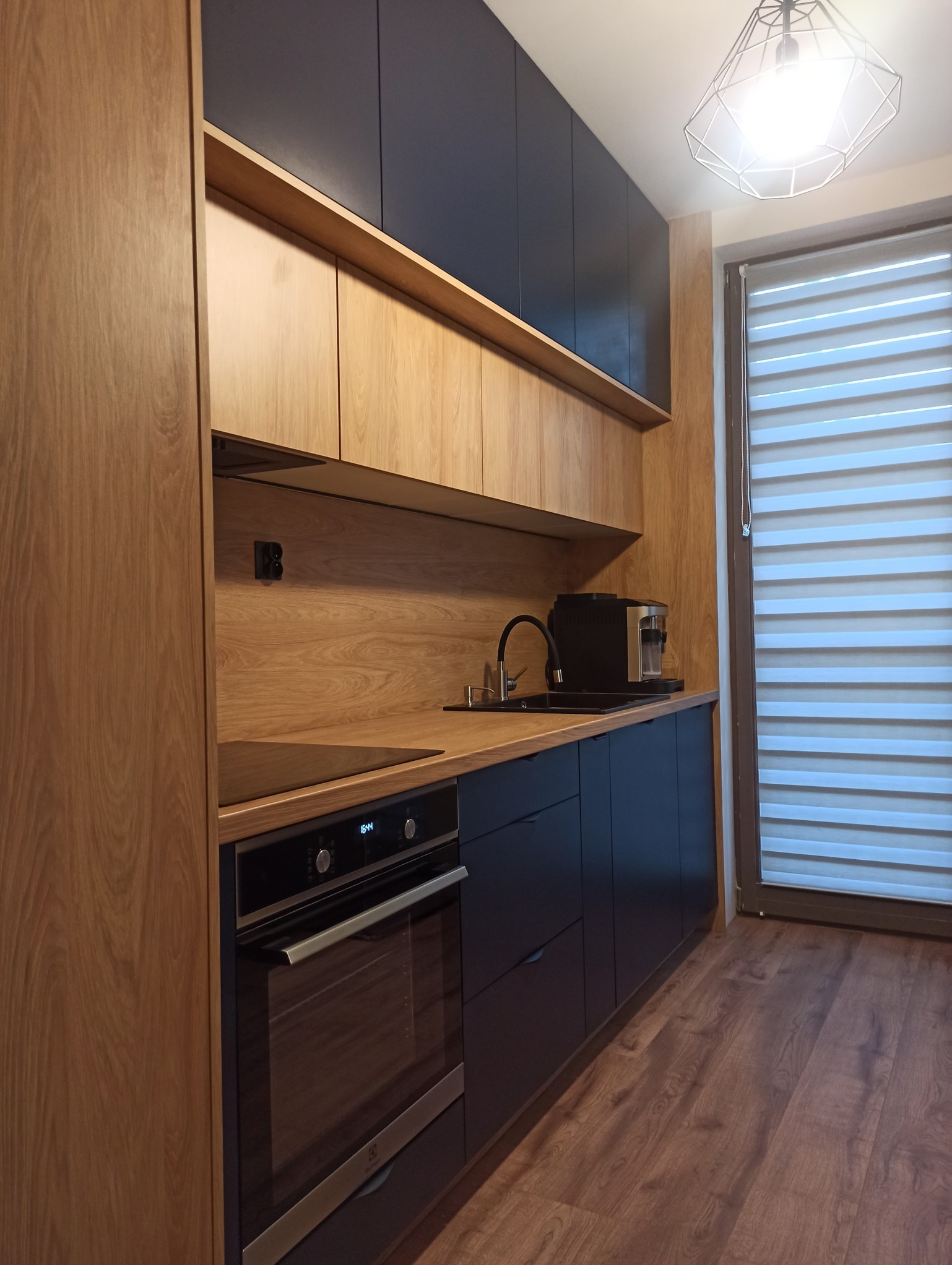 Nowoczesna kuchnia na wymiar z drewnianymi frontami i czarnymi szafkami wiszącymi, oświetlona lampą drucianą. Widoczny piekarnik i ekspres do kawy.