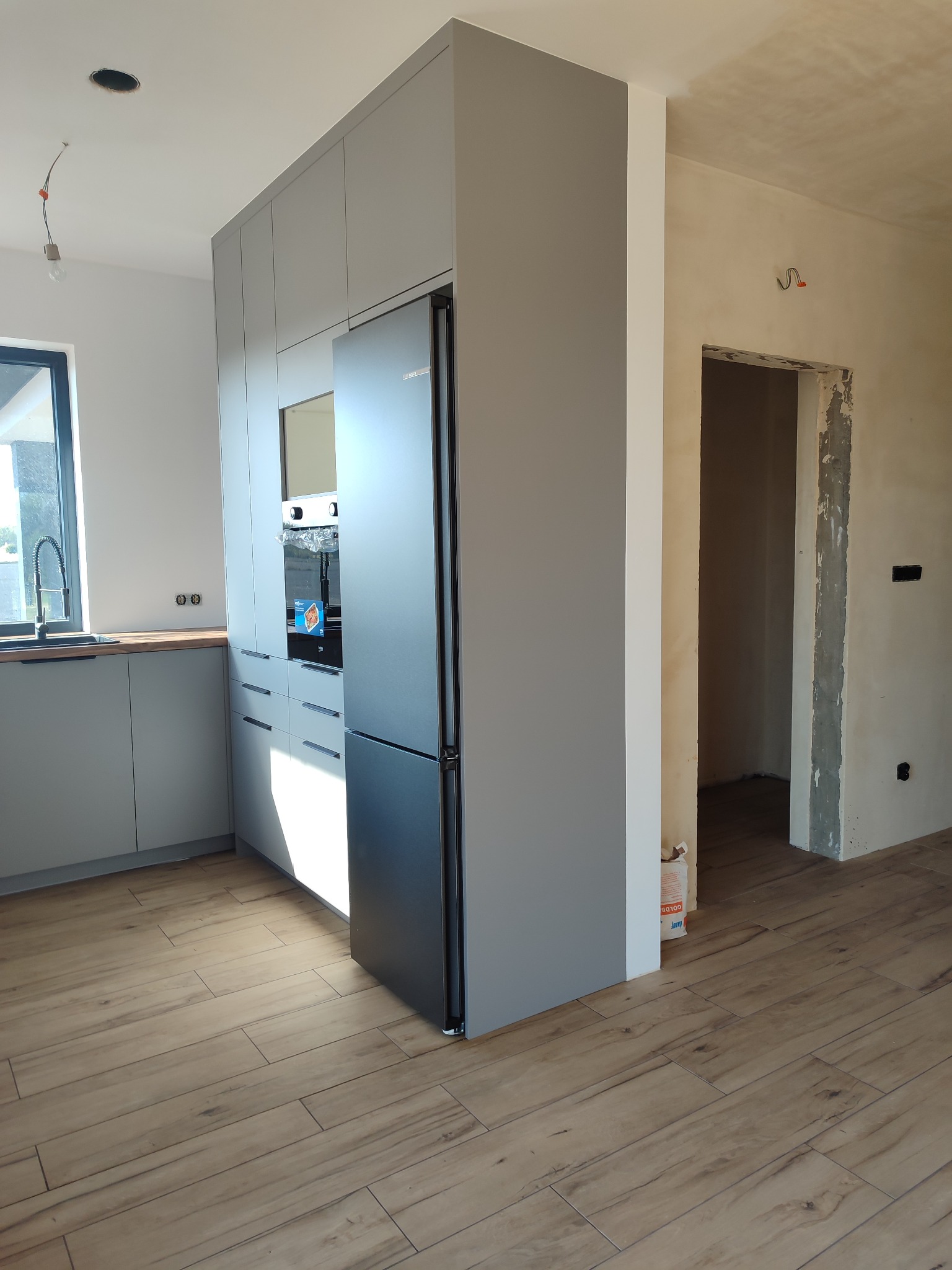 Szara zabudowa kuchenna z lodówką i piekarnikiem w surowym wnętrzu. Widoczne płytki podłogowe imitujące drewno i fragment ściany w stanie deweloperskim.