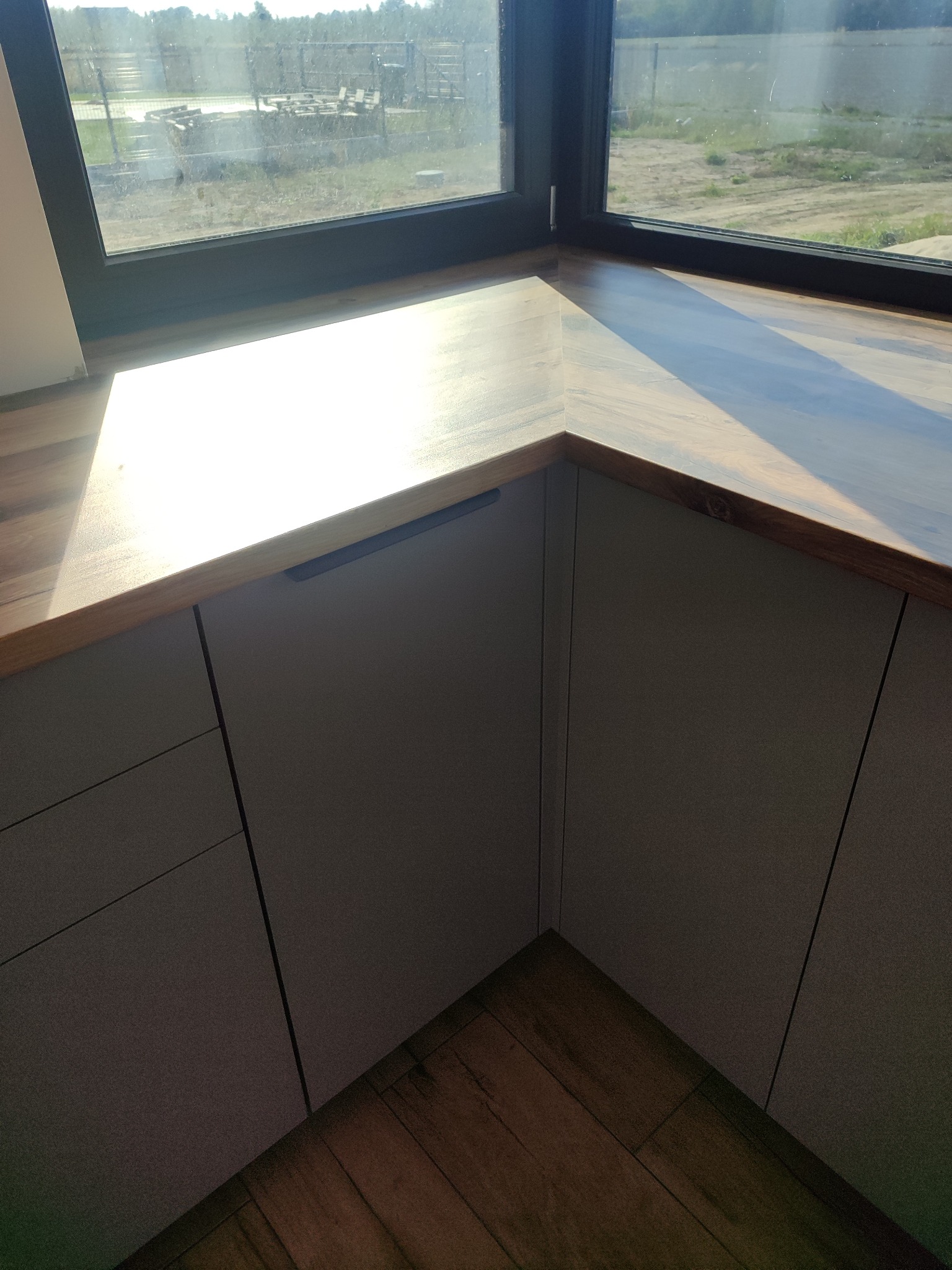 Szare szafki kuchenne z drewnianym blatem pod oknem, widok z góry, minimalistyczny design, drewniana podłoga, jasne światło słoneczne.
