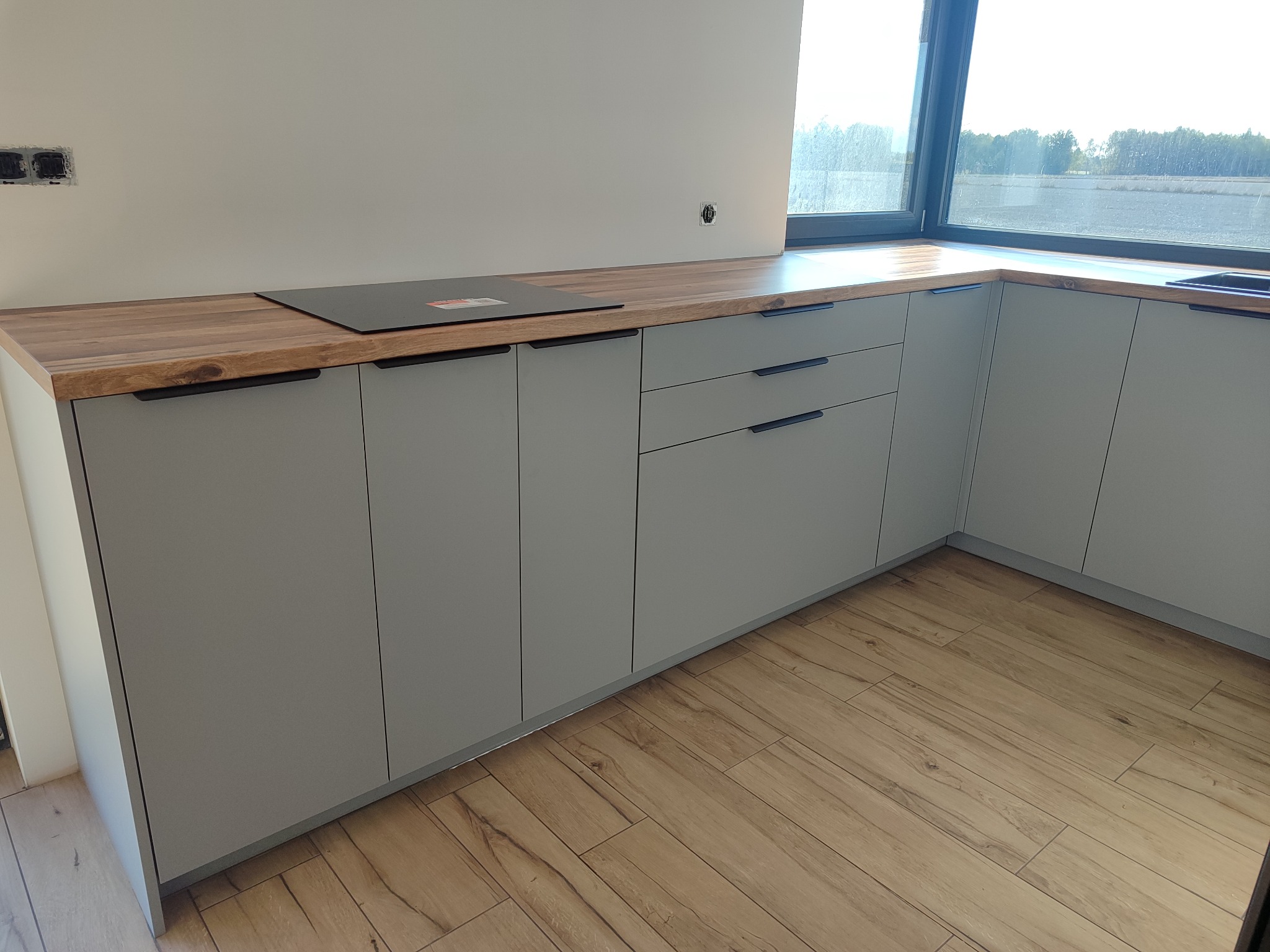 Szare, minimalistyczne szafki kuchenne z drewnianym blatem i czarnymi uchwytami, płyta indukcyjna, okno z widokiem na zewnątrz, drewnopodobna podłoga.