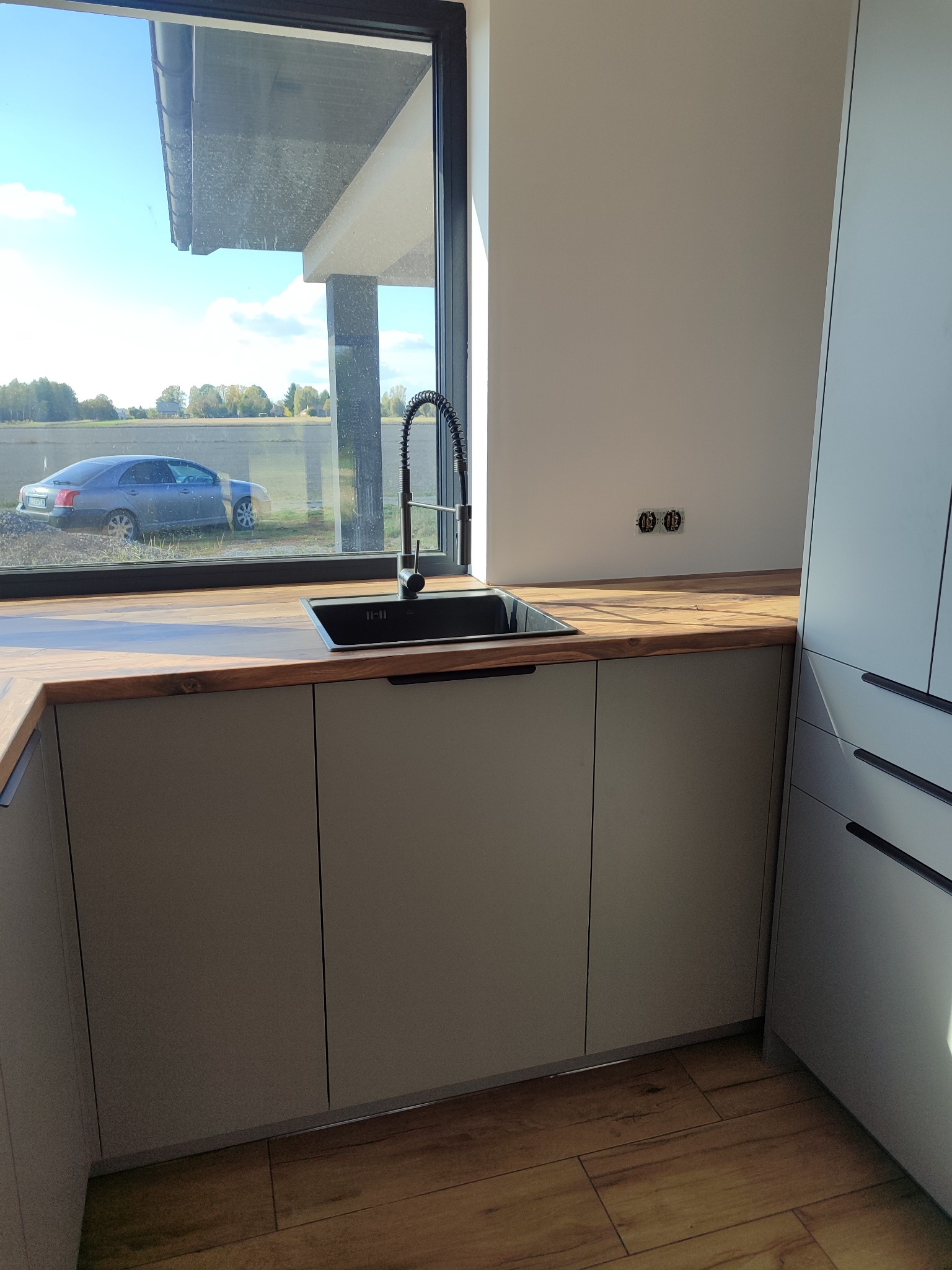 Nowoczesna kuchnia z szarymi szafkami, drewnianym blatem i czarnym zlewem. Za oknem widoczny samochód i zieleń. Minimalistyczny design z elementami natury.
