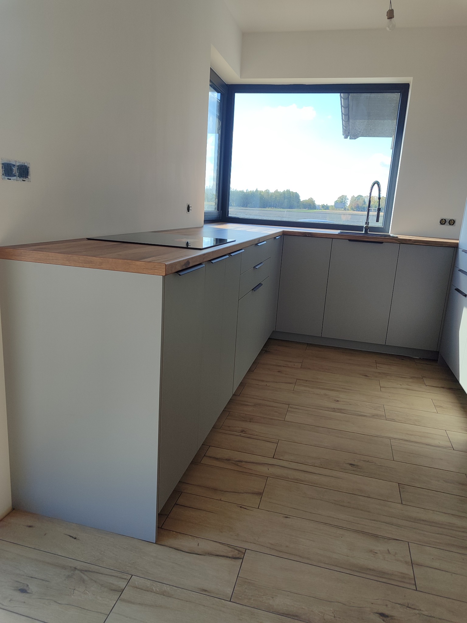 Szara, nowoczesna kuchnia z drewnianym blatem i dużym oknem z widokiem na zieleń. Minimalistyczne uchwyty, drewnopodobna podłoga. Widoczne gniazdka elektryczne.